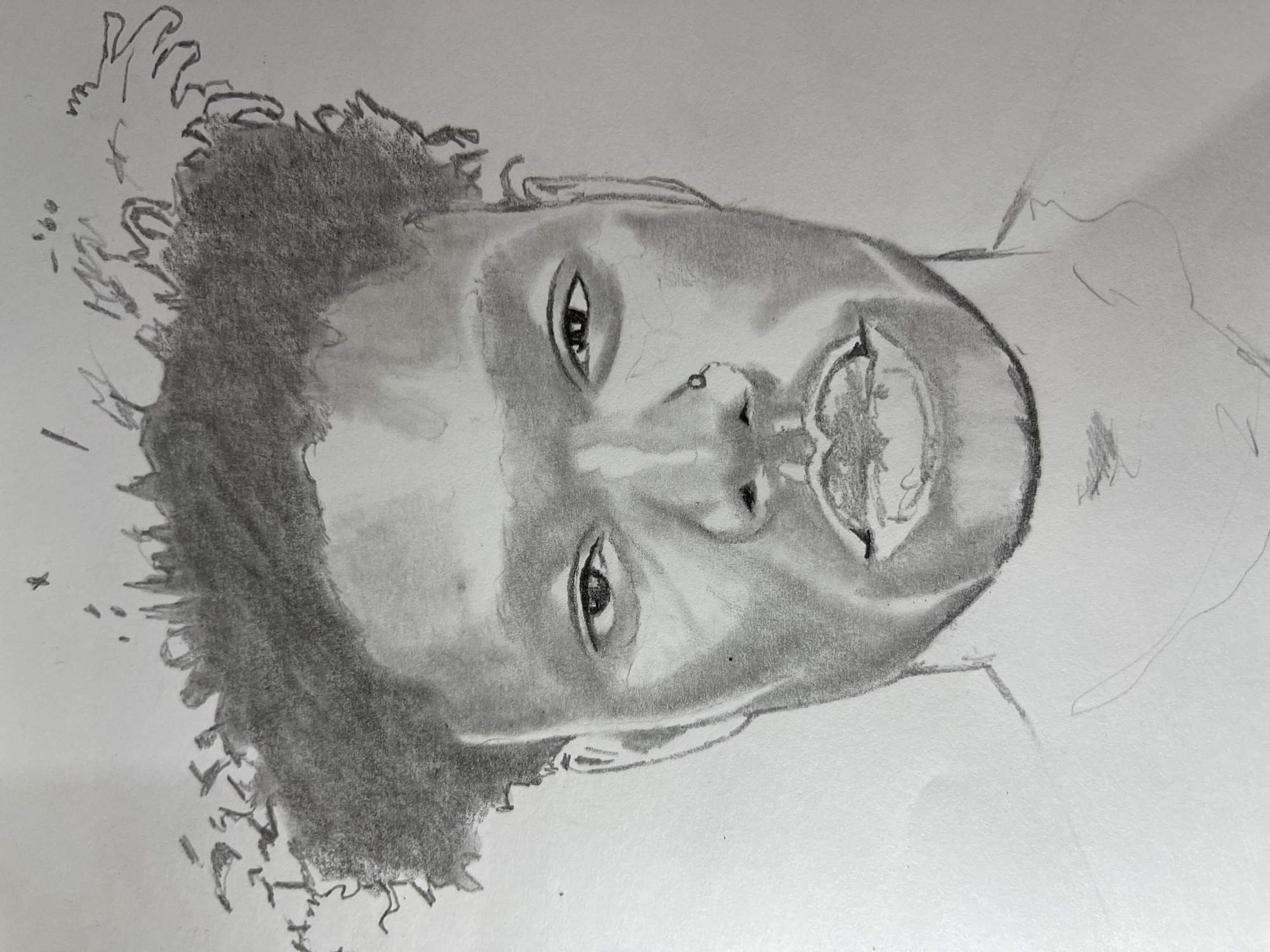 The image is a pencil portrait drawing on white paper, depicting the head and upper shoulders of a dark-skinned individual, likely male, centered and facing directly forward. The subject has short, curly, textured hair, open eyes looking straight ahead, and a slightly open mouth revealing some teeth. A small ring is visible in the subject's left nostril. The drawing utilizes various shading techniques to define facial contours, suggesting a light source illuminating the left side of the face and casting shadows on the right. The neck and shoulders are minimally outlined. The background is unadorned white paper, providing no environmental context. The drawing itself does not depict a location in the city of Jos, Nigeria.