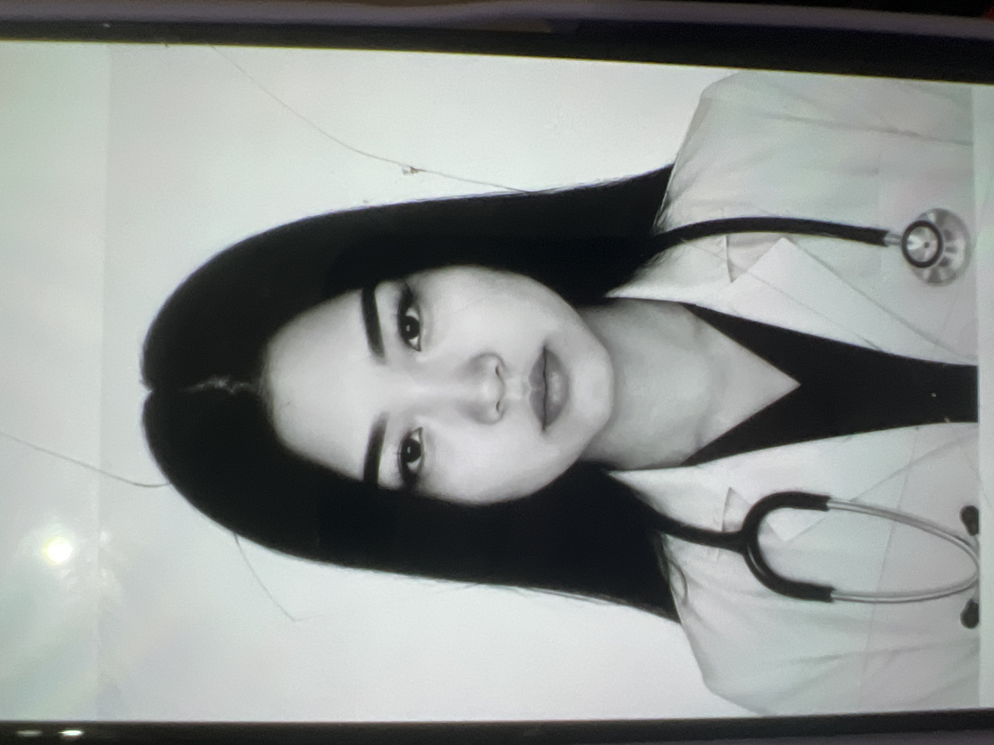 This is a black and white passport-style photograph of a young Asian woman wearing a white lab coat over a dark V-neck top. She has long, straight black hair parted in the middle. A stethoscope is draped around her neck, with the earpieces resting on her shoulders and the chestpiece visible on the right side of her coat. Her expression is neutral, and she is looking directly at the camera. The background is plain and light-colored, with some subtle smudges and a bright, circular light source visible in the upper left quadrant, suggesting artificial lighting. The image appears to be taken indoors. There is no visible indication of the city of Kara-Dıykan, Kyrgyzstan, in the image itself.