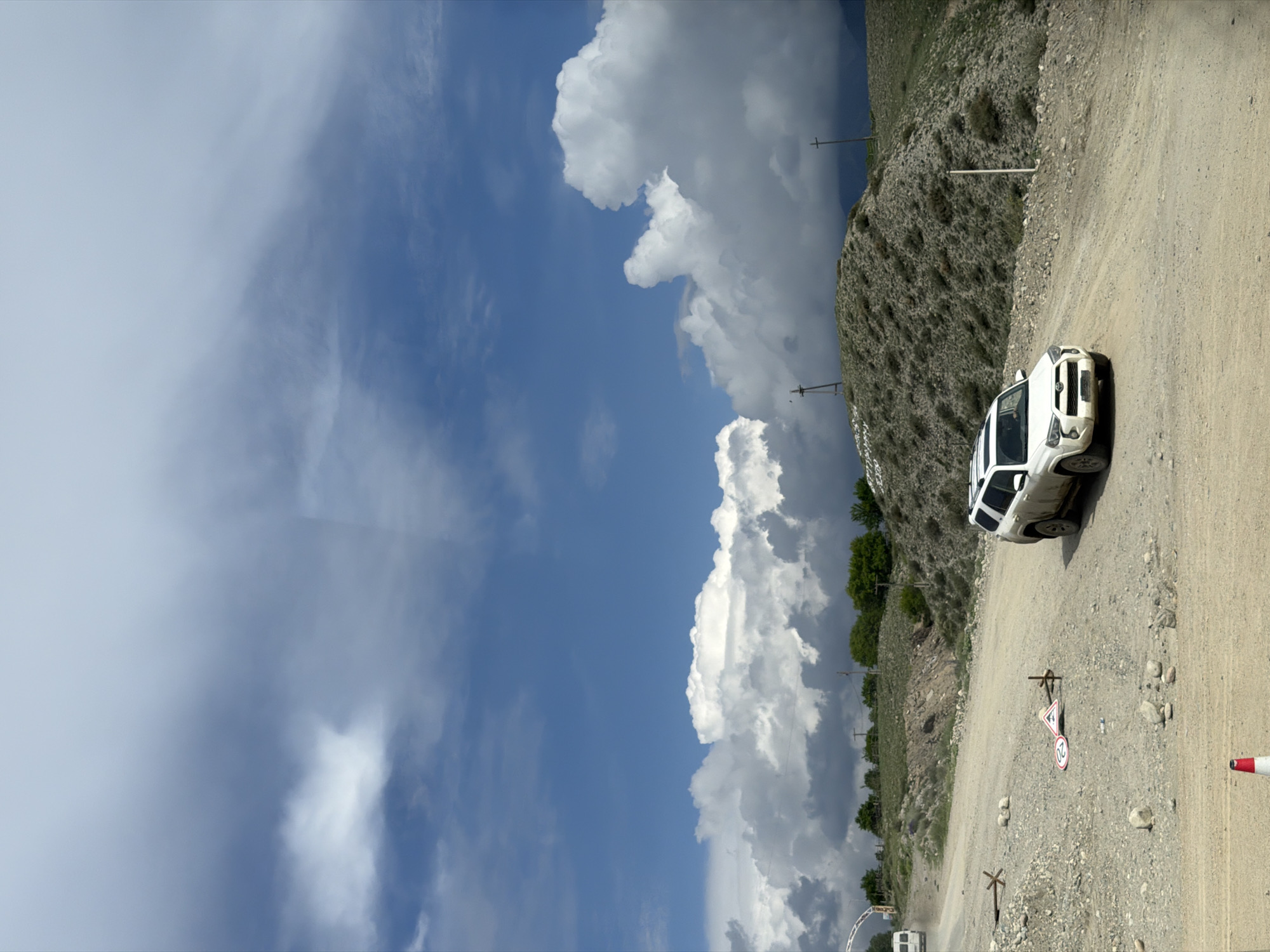 A white SUV is parked on a dirt road in a mountainous, rural setting. The car is positioned at an angle on the right side of the frame, with its front facing towards the left. The road is unpaved and appears to be made of gravel and dirt, with some larger rocks scattered along its surface.
The landscape is characterized by steep, rocky hillsides covered in sparse vegetation. A vast blue sky dominates the upper portion of the image, filled with dramatic, puffy white clouds, suggesting fair weather with potential for changes. The lighting indicates it is daytime, likely midday or early afternoon, given the brightness and the shadows.
There are several signs visible on the side of the road. One sign, partially obscured, shows a triangle with an exclamation mark, often indicating a warning. Below it, another circular sign with a red border displays the number "20", likely a speed limit. Another smaller sign with an "X" symbol is also present. Utility poles with wires run along the side of the road, indicating the presence of infrastructure.
In the distance, more hills and some scattered trees can be seen, with a hint of a valley or a wider expanse beyond. The overall impression is one of a remote, scenic location, possibly a popular route for off-road vehicles or hikers, given the road conditions and the presence of signs. No people are visible in the image, and the car appears to be stationary, suggesting it is either parked for a break or has stopped for a specific reason. The scene evokes a sense of adventure and the rugged beauty of the natural environment.