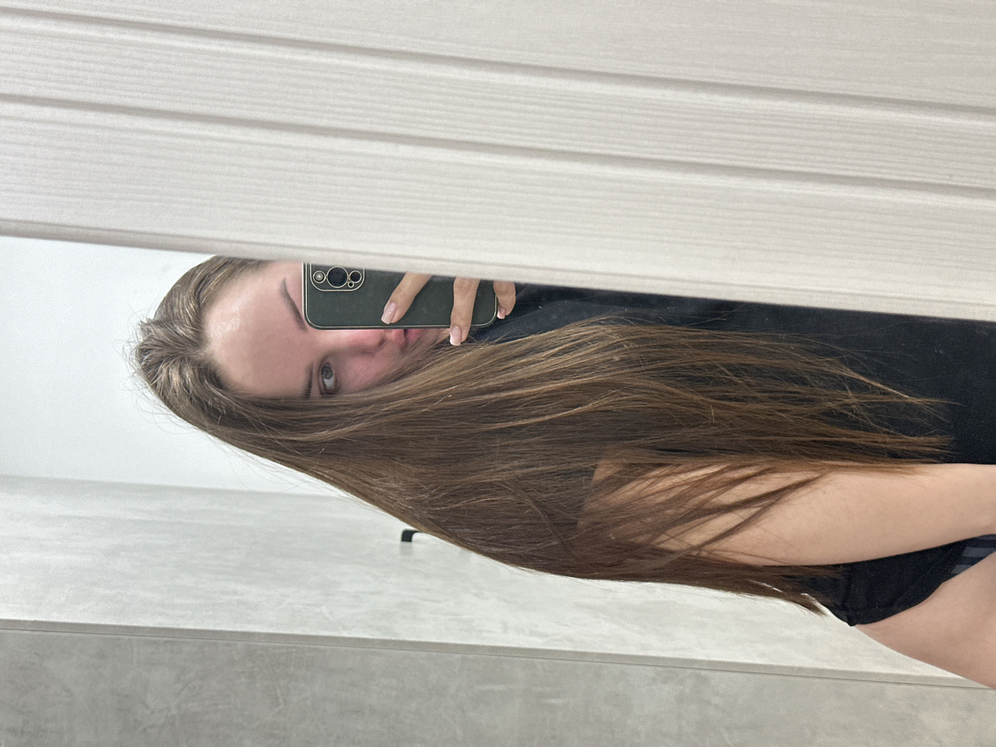 A woman is taking a selfie in a mirror. The reflection shows part of her face, long brown hair, an arm, and a black top. She's holding a smartphone with a green case. The mirror is mounted on a wall, and part of a white, paneled blind is visible above the mirror. The background consists of a light-colored wall and a grey surface. There is no indication of the location being Chaikovsky, Russia.