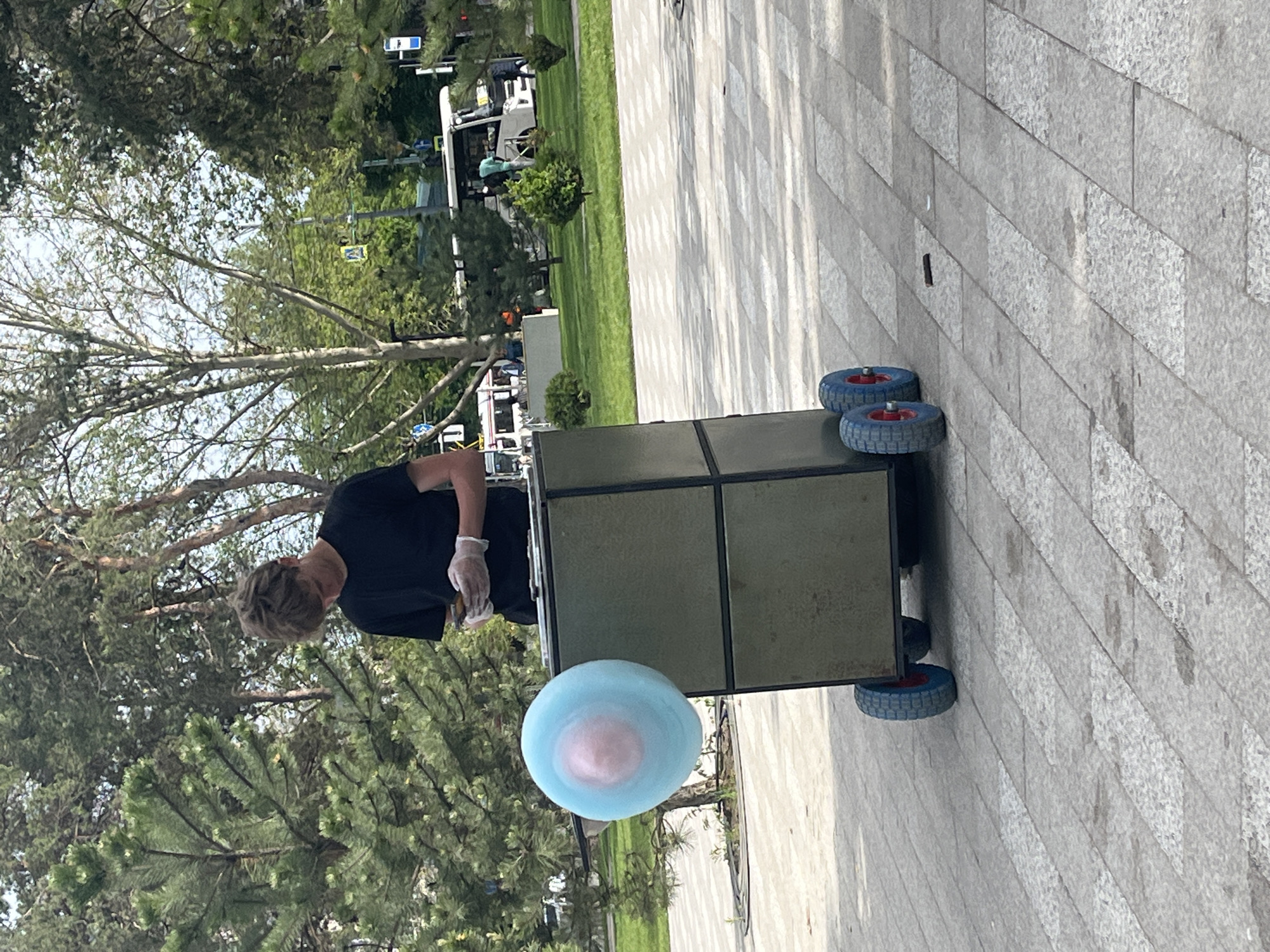A street vendor in Krasnodar, Russia, wearing gloves, is making cotton candy on a cart with blue wheels. The cart is located on a patterned, tiled sidewalk in a park area. The finished cotton candy is light blue and pink. Trees and greenery are visible in the background. In the distance, there is a white van.
