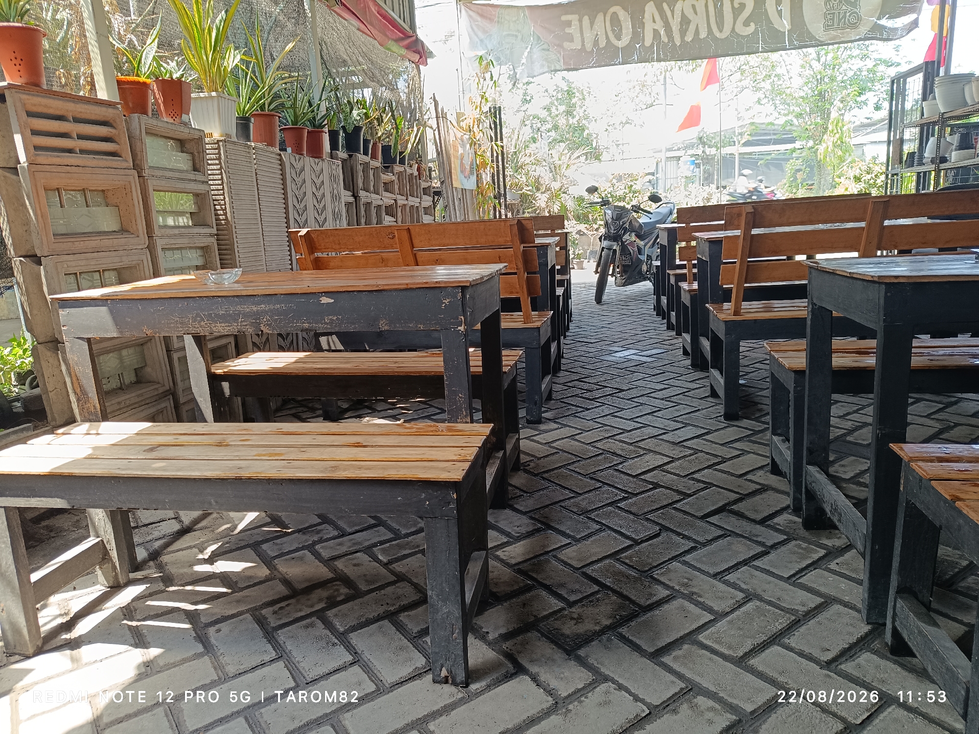 An outdoor seating area with wooden tables and benches is depicted. The tables and benches are dark-colored with wooden seating surfaces and tabletops. They are arranged on a patterned brick floor. In the background, there are stacks of decorative concrete blocks and potted plants. A motorcycle is parked to the right of the tables, and a banner with text is visible hanging overhead. The scene appears to be an outdoor cafe or dining establishment.