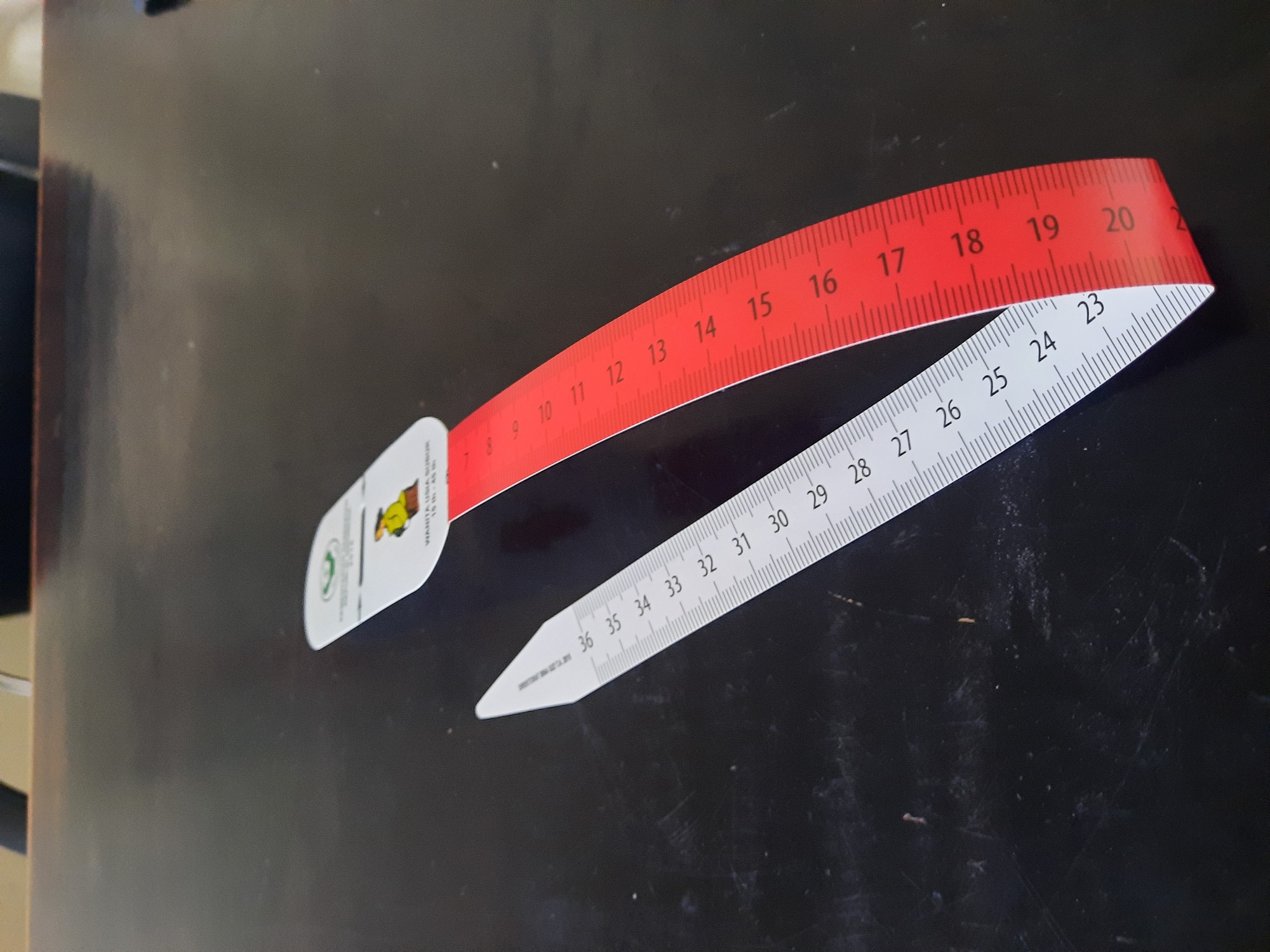 The image shows two measuring tapes on a dark, possibly wooden, surface. The left tape is white with black markings and numbers, showing measurements from 23 to 36, with smaller tick marks for finer increments. This tape is pointed at one end, like a dart. The right tape is red with white markings and numbers, appearing to be a continuation or a different part of a measuring tape. It shows numbers starting from 7 up to 20. A white label is attached to the red tape with some text and a small illustration of a person. The label text includes "WANITA USIA SUBUR" and "15 th - 45 th".
Based on the text "WANITA USIA SUBUR" which translates to "Women of Reproductive Age" and the context of measuring tapes, this image is likely related to a health program or initiative in Lhokseumawe, Indonesia, possibly for monitoring or measuring women of reproductive age. The dark surface could be a table in a health clinic, office, or community center within the city. The lighting appears to be overhead, casting some slight reflections on the surface. The image is captured from a slightly elevated angle looking down at the measuring tapes.