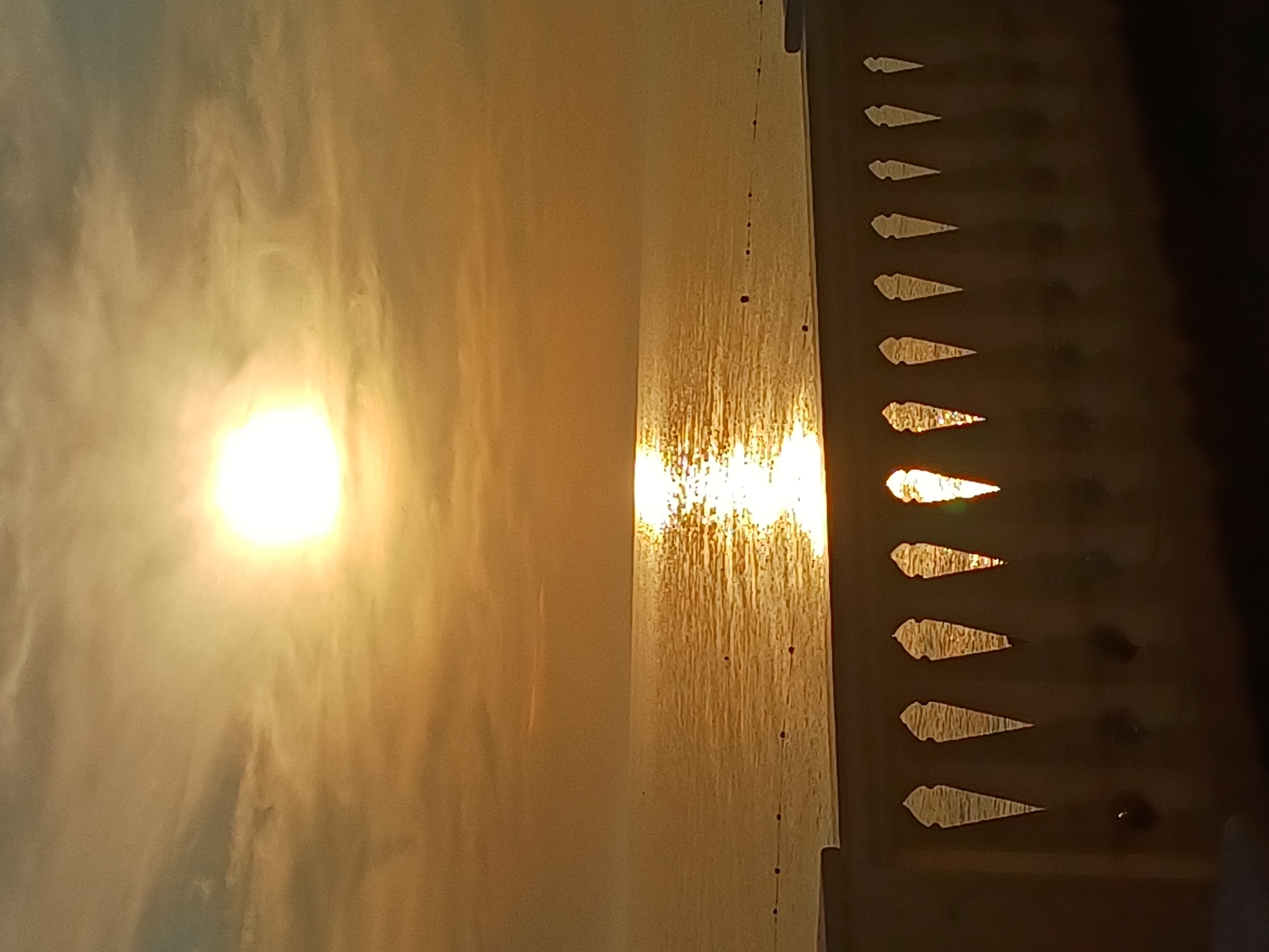 This forensic description details a serene coastal scene at either sunrise or sunset, characterized by warm, golden light. The sun, a bright, intense orb, is positioned in the upper center of the frame, partially veiled by wispy, orange-tinted clouds that dominate the upper sky. Its powerful rays cast a brilliant, shimmering path of light across the calm, dark-orange sea, extending from the horizon towards the viewer. The horizon line is flat and distant, separating the sky and the vast expanse of water.
In the middle ground, a line of dark, spherical buoys is visible on the water's surface, extending horizontally across the frame, suggesting a demarcated area within the sea. The foreground is dominated by a dark, silhouetted architectural feature, specifically a balustrade or railing, with a repeating pattern of decorative, pointed, keyhole-shaped openings. These openings partially frame and reflect the shimmering sunlight from the sea, creating bright, illuminated patterns within their dark forms. The surface supporting the balustrade is a dark, undefined mass. No individuals or specific human activity are discernible in the image. The scene is located in the city of Sochi, Russia.
