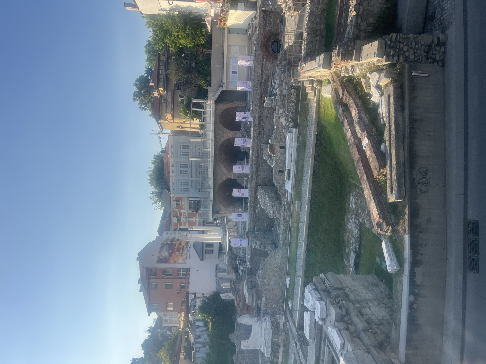 The image shows the ruins of the Roman Stadium of Philippopolis in Plovdiv, Bulgaria, on a bright, clear day. The ancient stadium's stone seating and remnants of walls are visible, with patches of green grass growing among the ruins. In the background, modern buildings rise above the ancient site, including a tall, orange-hued building with a large mural on its side. Several white banners with text are displayed on poles within the archaeological site, likely providing information to visitors. The sky is a clear, pale blue.