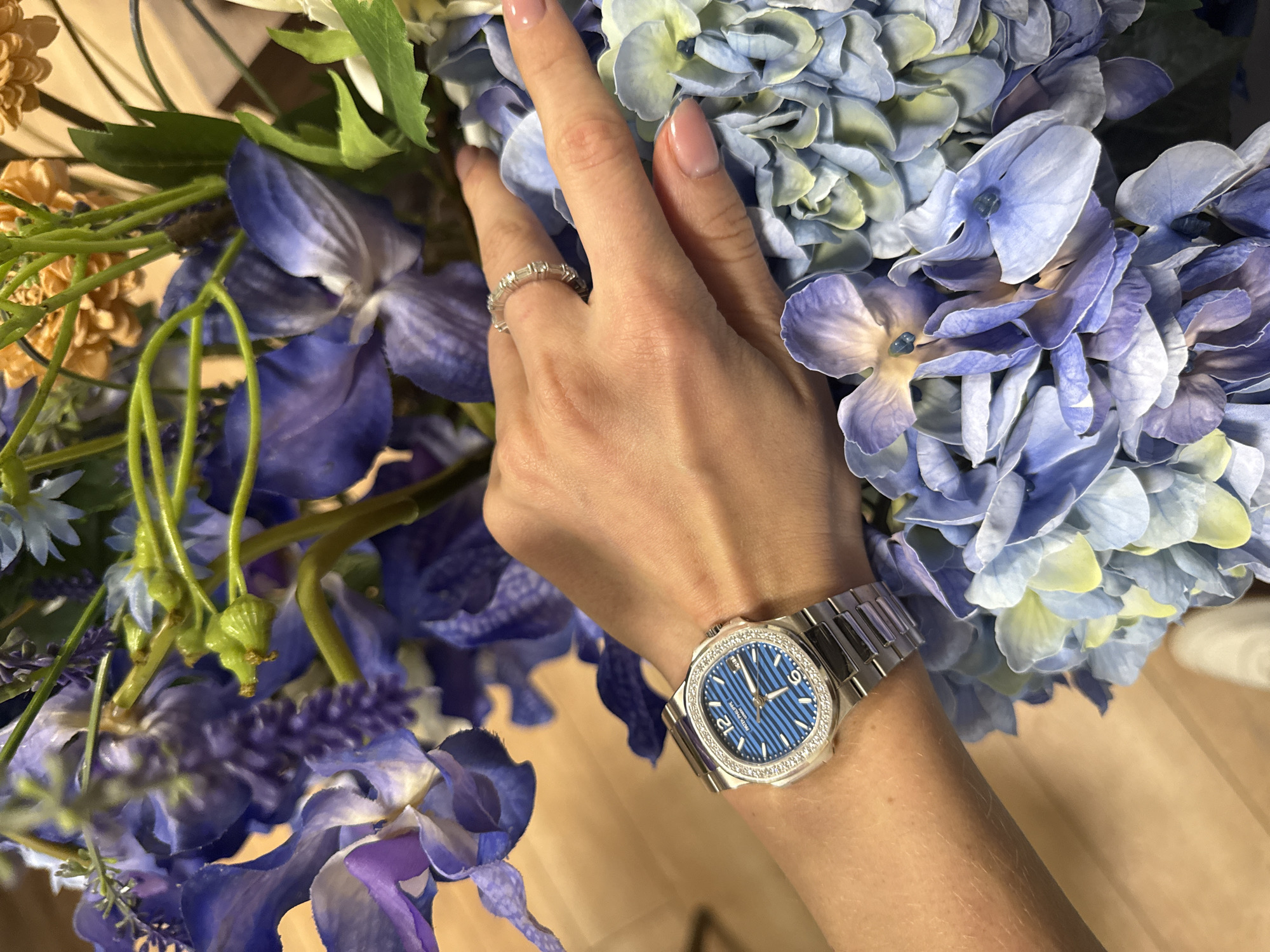 A woman's hand, adorned with a diamond ring and a blue-faced wristwatch with a silver band and diamond bezel, rests amidst a bouquet of blue and white flowers. The scene is indoors. There is no evidence to support a location in Moscow, Russia.
