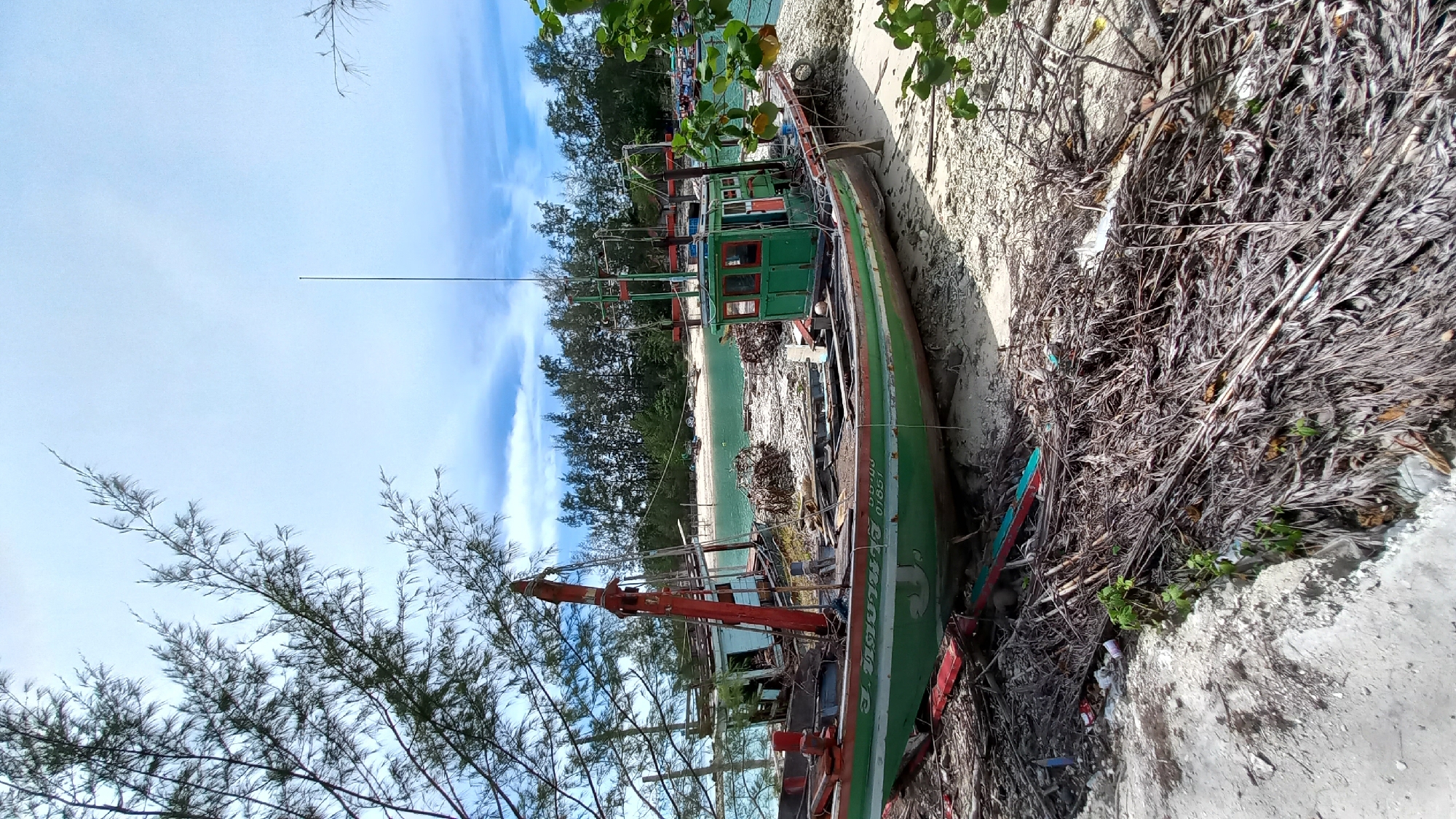 In the coastal area of Tambon Ko Pha Ngan, Thailand, a weathered green fishing boat rests on a sandy shore, partially surrounded by debris. The boat exhibits signs of age and disuse, with a visible inscription in Thai script and a registration number "01651" on its hull. To the left of the main boat, another partially dismantled vessel, featuring a prominent red crane, is visible. The background reveals a serene beach with lush green trees lining the horizon and calm, turquoise water. The sky above is a mix of pale blue with scattered white clouds. The overall scene depicts a quiet, possibly abandoned, marine area.