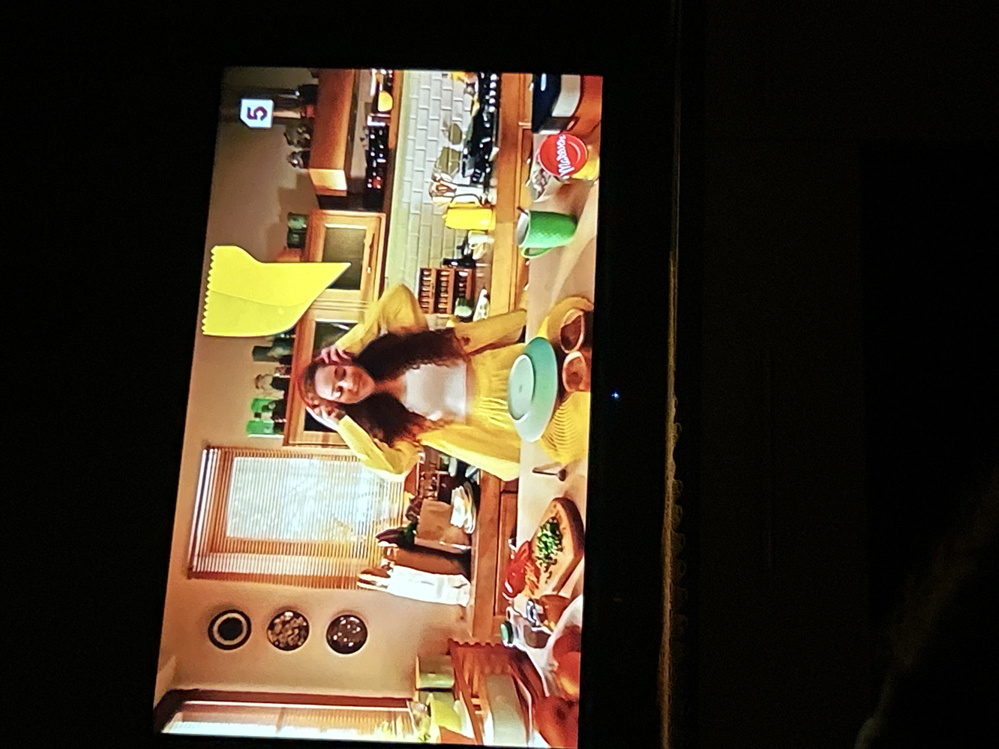 The image shows a kitchen scene on a television screen. A young woman wearing a yellow jacket and skirt stands in the center, with her hands near her head. She is looking down, and her expression is partially obscured. In front of her on the counter is a cutting board with chopped vegetables, a red pepper, and jars. To the right of the cutting board are a green mug, a bowl with contents, and a red and white circular tin. Behind her, shelves hold various items, and a yellow object, possibly a placemat, hangs on the wall. A window with blinds is visible in the background. The television is displaying this image in a dark room. There is no information in the image to suggest any connection to the city of Gelendzhik, Russia.