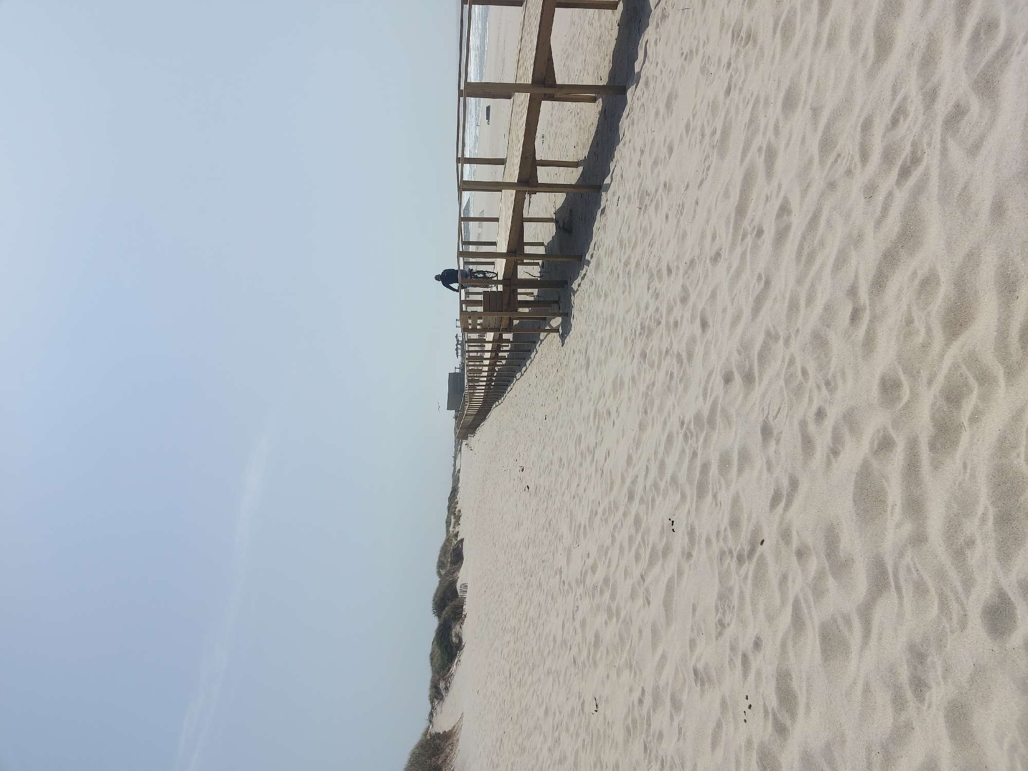 The image depicts a beach scene in Gafanha da Nazaré, Portugal. The view is oriented vertically, showing a wide expanse of light-colored sand. A wooden boardwalk runs along the edge of the beach, with a person riding a bicycle on it. The sky is clear and pale blue. Vegetation is visible along the lower left part of the image, suggesting dunes.