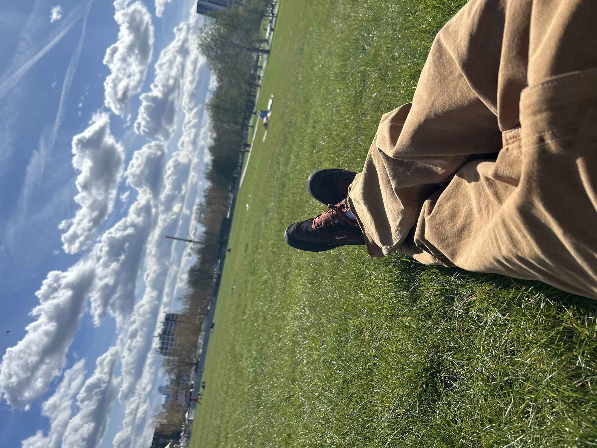 A low-angle, slightly tilted, outdoor shot depicts a person sitting on a lush green lawn on a sunny day, partially obscuring the surrounding environment. The person is wearing tan-colored, corduroy pants, and brown and black Nike shoes with rust-colored laces. The person's legs are bent at the knees and drawn up toward the torso. A patch of grass is visible between the person's shoes and pants.
In the background, a wide expanse of green lawn stretches toward a line of trees and buildings in the distance. There appear to be other individuals lying on the grass further away. The sky is bright blue, filled with large, puffy white clouds. Contrails are visible near the top left of the frame. Based on the cityscape visible and features of the park, the location is likely in Hyde Park, London, United Kingdom.