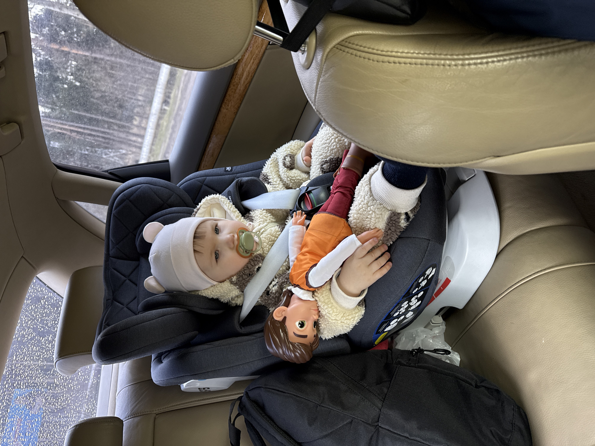 A young child sits in a car seat in the back of a vehicle, wearing a light-colored hooded jacket and a hat with bear ears. The child has a pacifier in its mouth and is holding a doll. A black bag sits at the base of the car seat, and the vehicle's interior is beige. The outside world is visible through the car window. The location in the city of Krapivka, Russia is unable to be verified as there is no additional information provided.