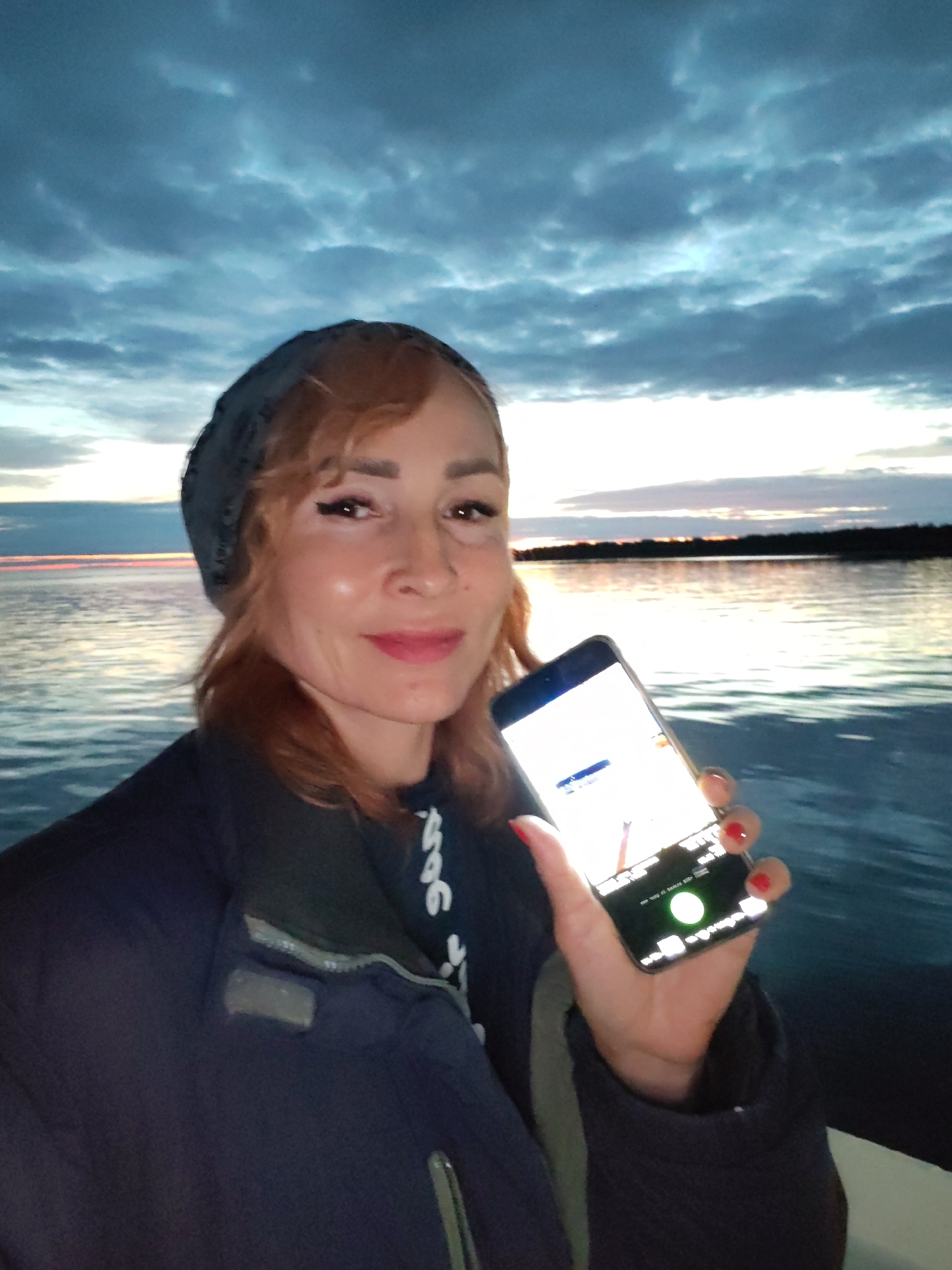 A woman with auburn hair, wearing a dark knit cap and a dark jacket, is taking a selfie in front of a body of water under a cloudy sky during what appears to be twilight. She is holding up a smartphone in her right hand, which displays a bright screen with some graphics. The water behind her is calm, with a hint of a sunset or sunrise on the horizon. The location is identified as Shcheleiki, Russia.