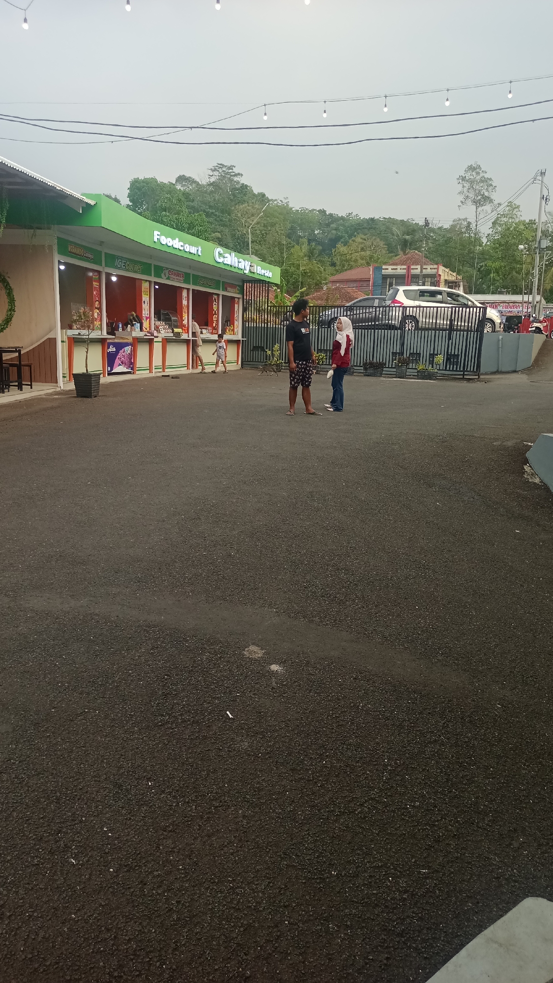 In Pakemitan, Indonesia, a food court named "Cahaya Resto" is visible. The food court features a green roof and several stalls with orange and white awnings. Various food items and drinks are displayed inside the stalls. Outside the food court, a black metal fence encloses a parking area where several cars are parked. Two individuals are standing in front of the fence. One is a man wearing a black t-shirt and patterned shorts, and the other is a woman wearing a red hijab and a white shirt. A young child is walking near the food court stalls. The ground is paved with asphalt. In the background, lush green hills and trees can be seen under a cloudy sky, suggesting a rural or semi-rural setting. The overall scene depicts a casual dining and gathering space.
