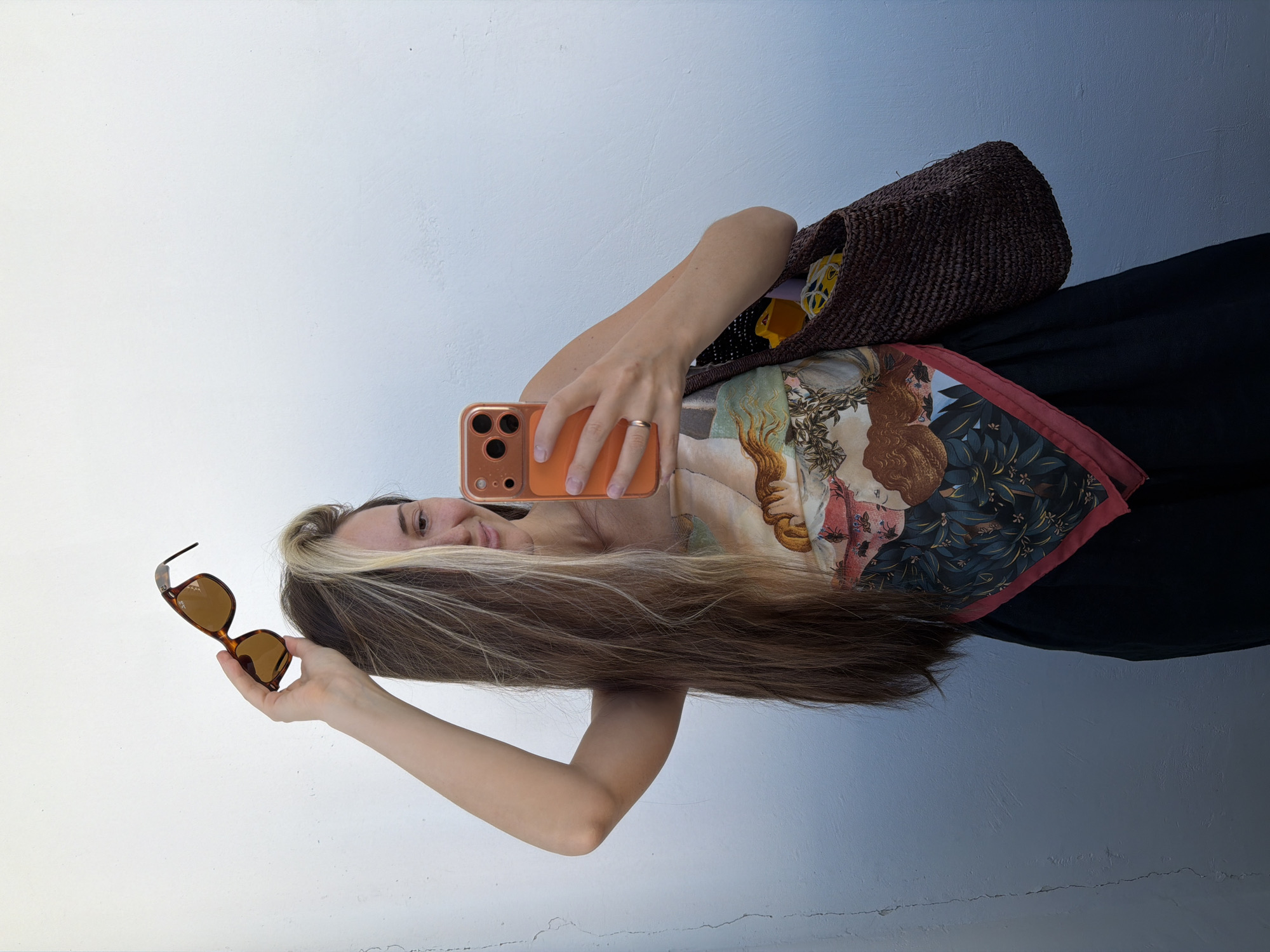 A young woman with long, blonde-streaked brown hair is standing in front of a white wall. She is holding a pair of sunglasses above her head with her right hand, while her left hand holds an orange-cased smartphone, as if taking a selfie. She is wearing a black dress with a colorful patterned scarf tied around her waist, featuring a depiction of classical figures and botanical elements. A woven brown bag is slung over her left shoulder, with a yellow object visible inside. The wall behind her has a gradient of white at the top to a light blue at the bottom, with a vertical crack on the left side. The image is taken outdoors, with natural lighting. No specific location in Khimki, Russia can be determined from the visual information provided.