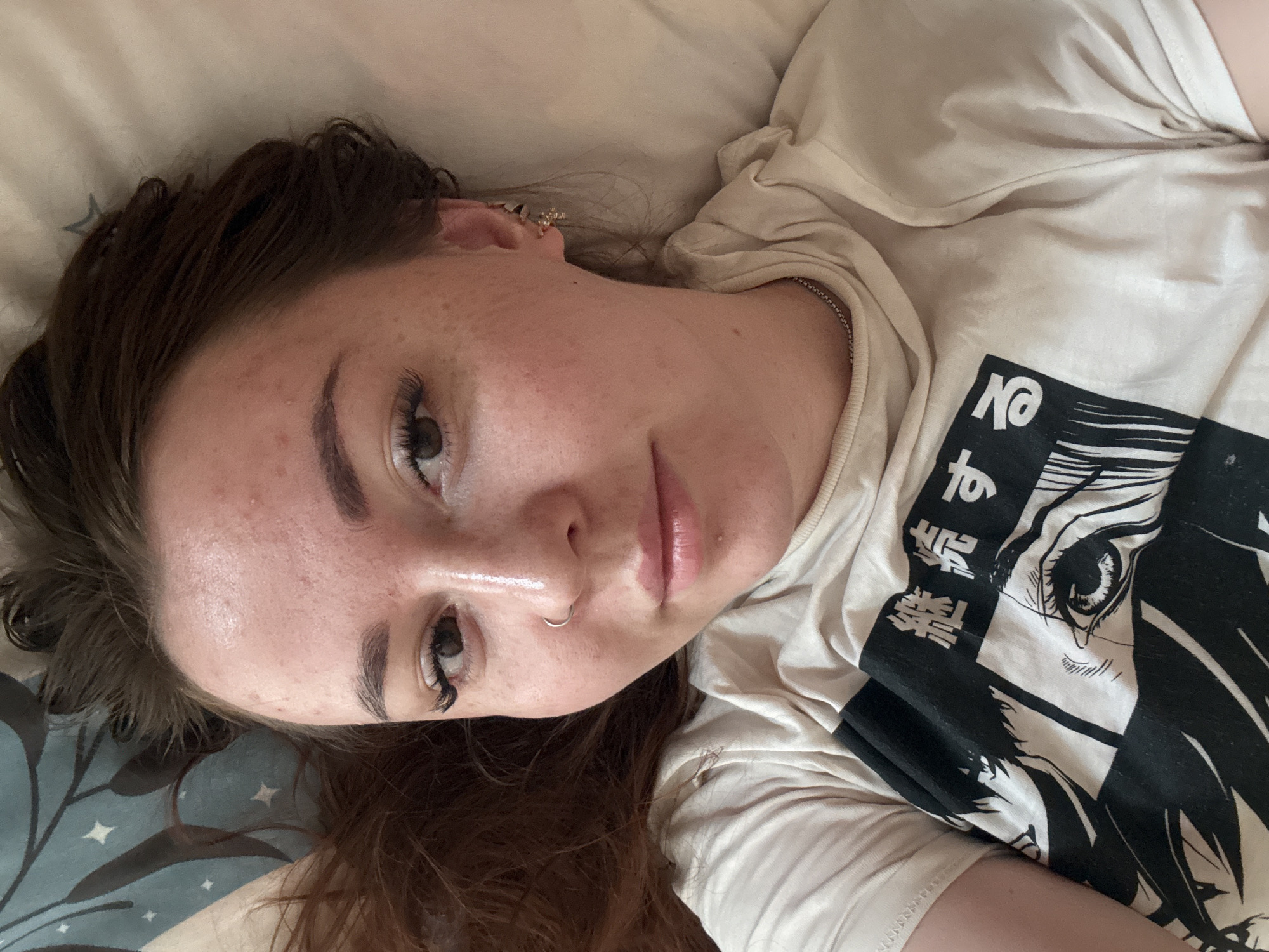 A young woman lies on a bed, seen from an above, slightly angled perspective. She is wearing a light-colored T-shirt with a black and white anime-style graphic. She has light skin, brown hair, and a nose ring. The bed has light-colored sheets, with a pillow in the lower left corner that has a blue and white patterned cover. The location of the scene is not apparent from the information provided.