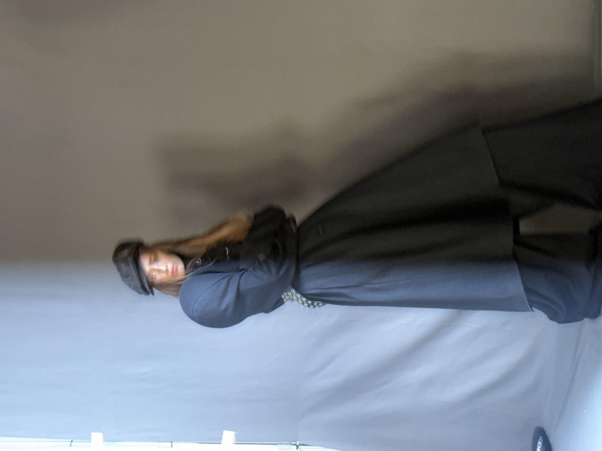 A woman with long brown hair and a flat cap is wearing a dark blue or black coat and skirt. She is standing in front of a gray background. The location is Unknown, Unknown.