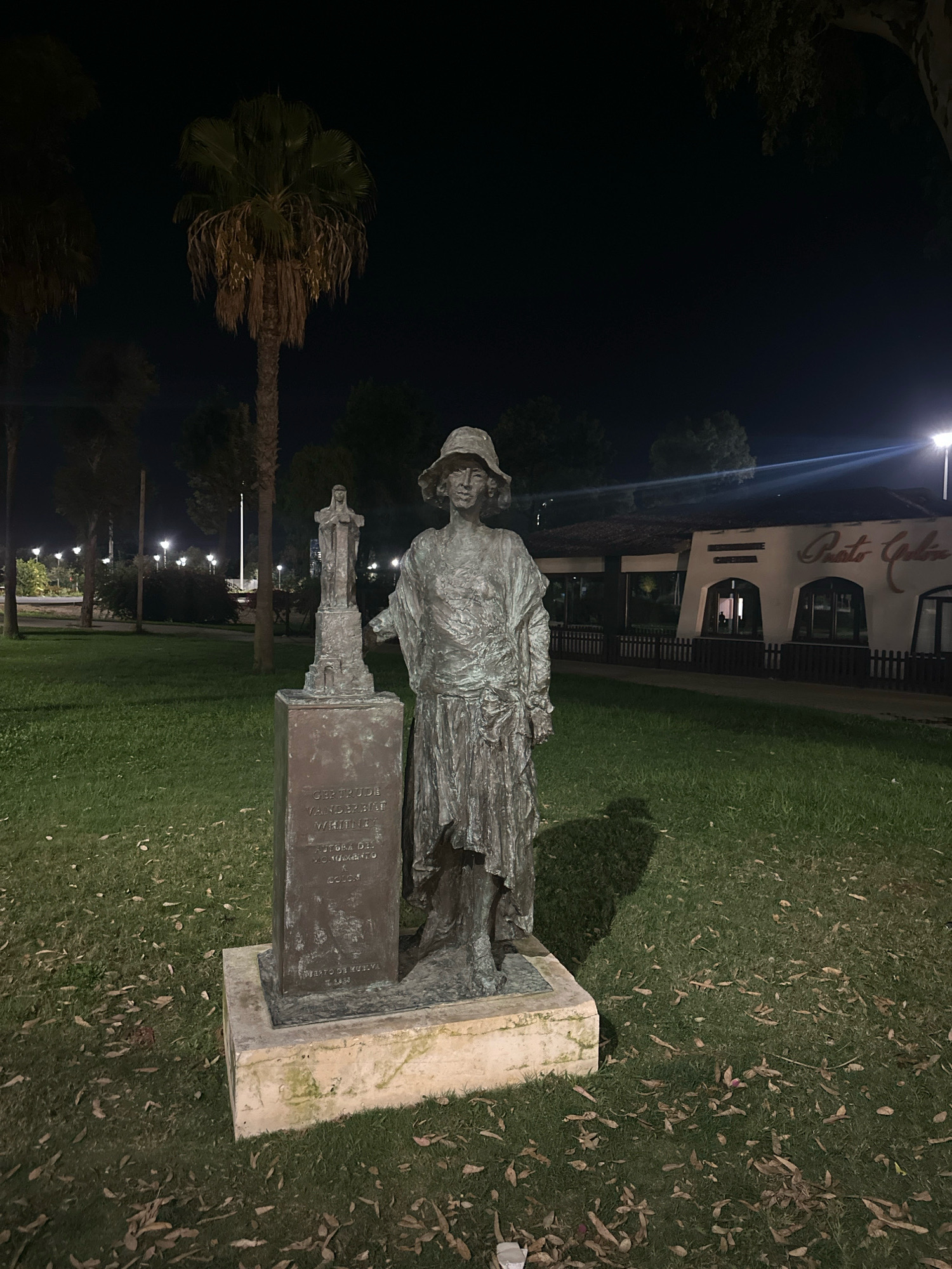 The image shows a bronze statue of a woman in a hat and long dress, standing next to a smaller statue on a pedestal. The pedestal has an inscription that reads "GERTRUDE VANDERBILT WHITNEY AUTORA DEL MONUMENTO X COLÓN." The statue is located in a park-like setting with grass, fallen leaves, and palm trees. In the background, there is a building with a sign that says "Puerto Colón." The scene is illuminated by streetlights, creating a nighttime atmosphere. This statue is located in the Puerto Colón area of the city of Huelva, Spain.