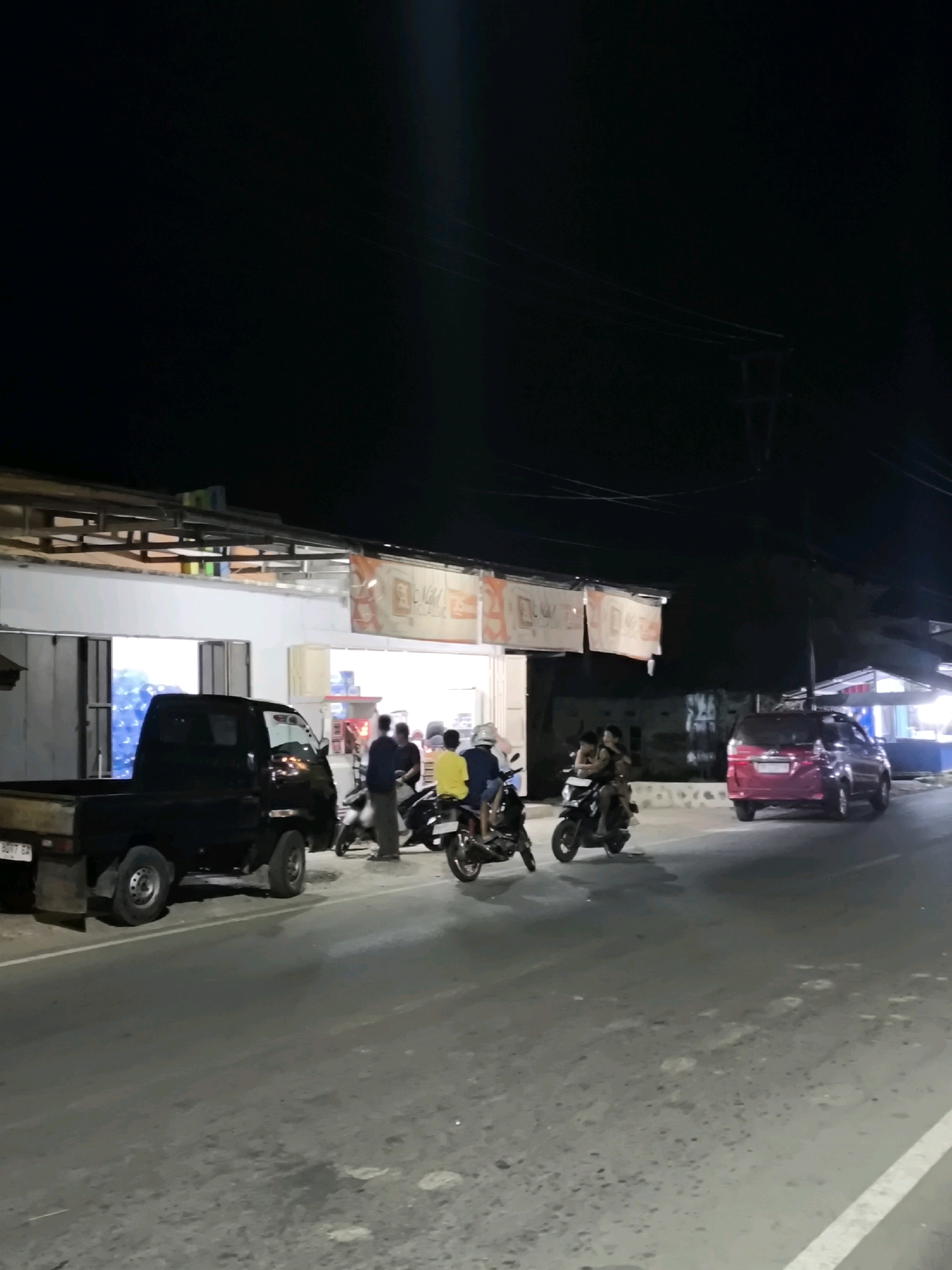 At night, a black pickup truck is parked on the side of a road in Moluo, Indonesia. A group of people are standing and sitting near a building with a storefront. Several motorcycles are parked or being ridden by individuals. A pink vehicle is parked further down the road. The scene appears to be a commercial area at night, with the building's interior illuminated.