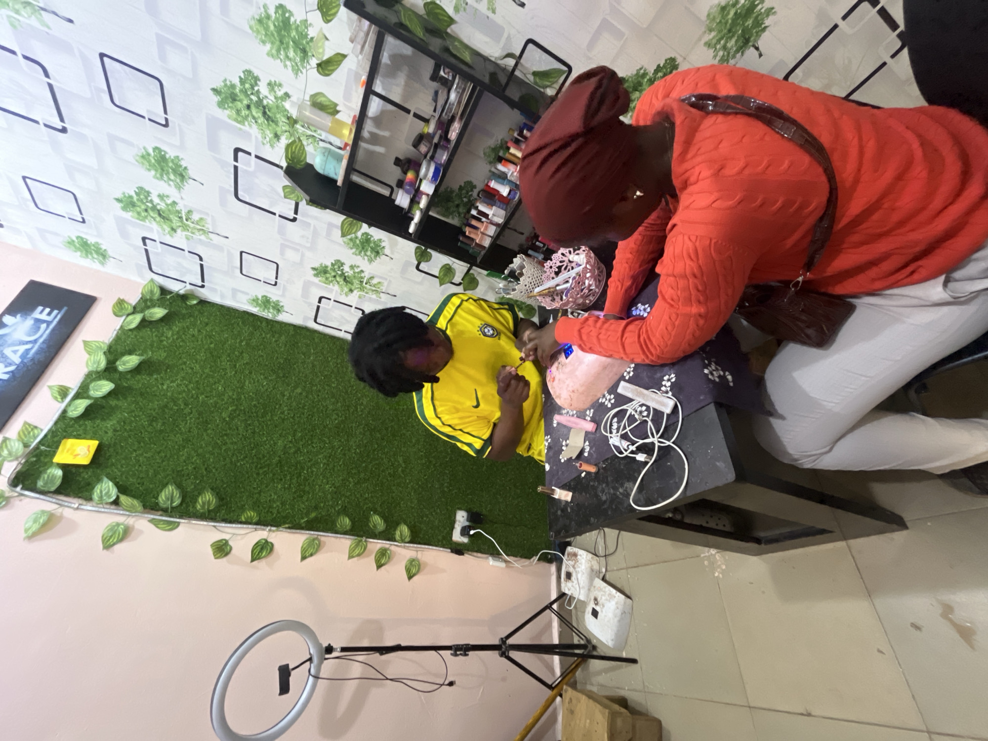 The image depicts a nail salon in Jos, Nigeria, where two individuals are engaged in a nail service. In the foreground, a person wearing a red, cable-knit sweater and a burgundy headscarf is seated, with their back to the camera, facing a black table. They appear to be receiving a nail treatment, as their hands are positioned over a pink acrylic nail form. A basket containing nail tools and a pink object are on the table, along with several nail polish bottles and some tangled wires.
Behind the table, another person, wearing a yellow and green soccer jersey, is seated and actively working on the recipient's nails, holding a tool with their right hand. This individual has dark hair styled in locs.
The environment is decorated with artificial greenery, including a wall covered in green turf and ivy vines draped along a light pink wall and a patterned wallpaper featuring green trees within white squares. To the left of the scene, a ring light on a tripod is set up, suggesting the potential for content creation. A black shelf unit filled with various nail products, such as polishes, creams, and brushes, stands against the patterned wall. The floor appears to be tiled, with some white material scattered near the legs of the table. The overall impression is a small, functional nail salon space.
