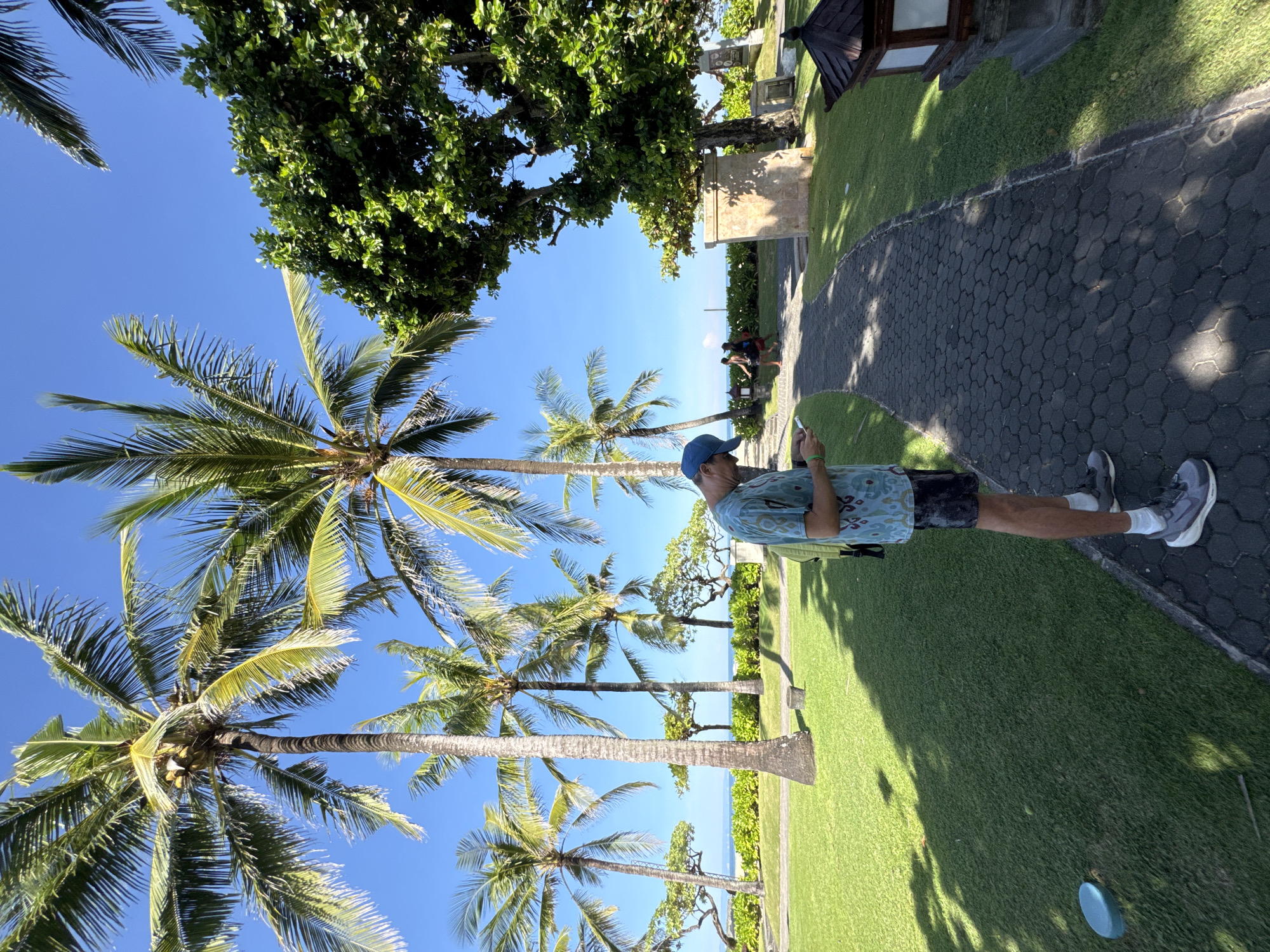 A daytime shot of a man standing on a brick pathway adjacent to a green lawn, with a view of palm trees against a blue sky, taken in Kuta Selatan, Indonesia. The man, wearing a blue cap, patterned short-sleeved shirt, dark shorts, and gray sneakers, is holding a phone and looking at it. The pathway runs alongside a manicured lawn, with palm trees visible in the background. The sky is clear and blue. Some buildings, trees, and a path with people are in the background on the other side of the lawn. A blue object is on the lawn near the bottom of the frame.