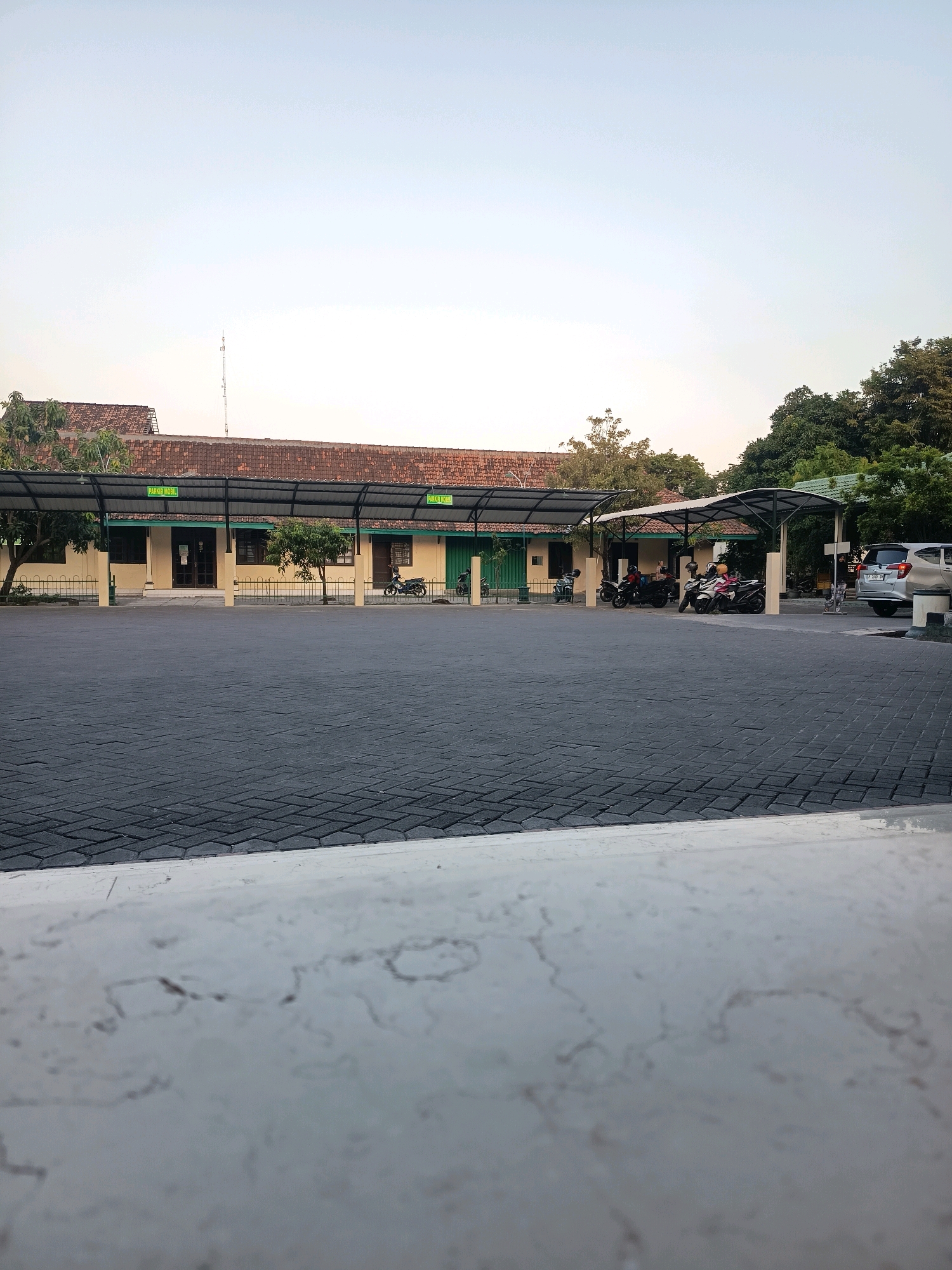 The image depicts an outdoor scene, likely a parking area, in Purwodadi, Indonesia. In the background, a long, single-story building with a tiled roof and cream-colored walls is visible. The building has several windows and doors, and a covered walkway runs along its front. Signs reading "PARKIR MOBIL" (Car Parking) are displayed above the walkway. Several motorcycles are parked under the covered area, and some people are seated on them. A silver SUV is parked to the right of the building, and a portion of another car is visible next to it. The foreground is dominated by a dark gray paver stone parking lot. Trees are scattered throughout the scene, with a denser cluster of trees on the right side. The sky is clear and pale blue, suggesting it is either early morning or late afternoon. The image is captured from a low angle, with a white, textured ledge or barrier in the immediate foreground.