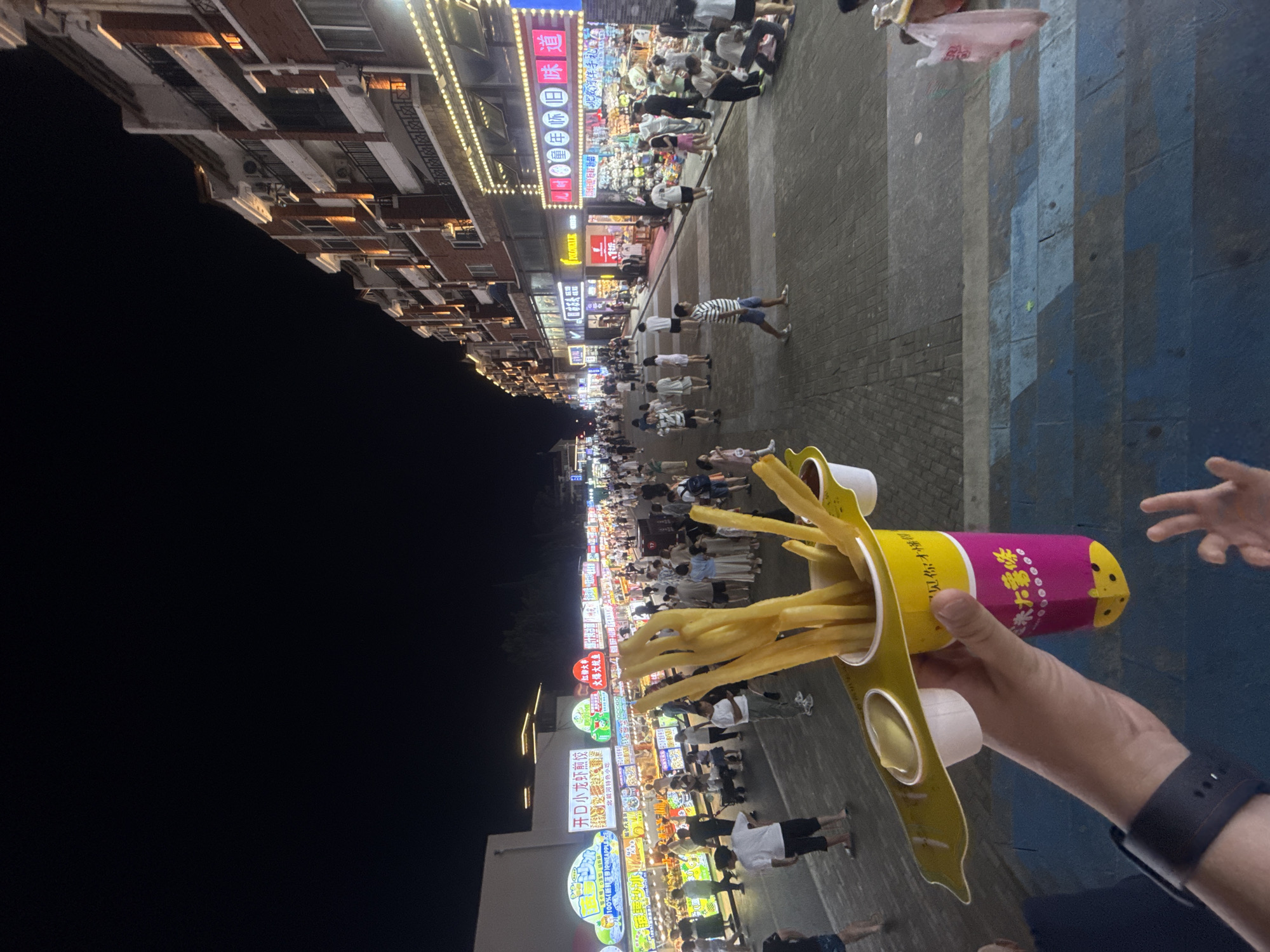 This is a first-person view of a person holding a fast-food cup with french fries and dipping sauces, in what appears to be a busy night market or pedestrian street in Qinhuangdao, China. The market is lined with shops and food stalls illuminated by bright lights. People are walking and browsing. In the foreground, the hand holding the french fries cup is visible. The background shows a street scene with buildings, more stalls, and a crowd of people. The architecture of the buildings suggests a modern commercial area. The overall atmosphere is lively and vibrant, typical of a bustling night market.