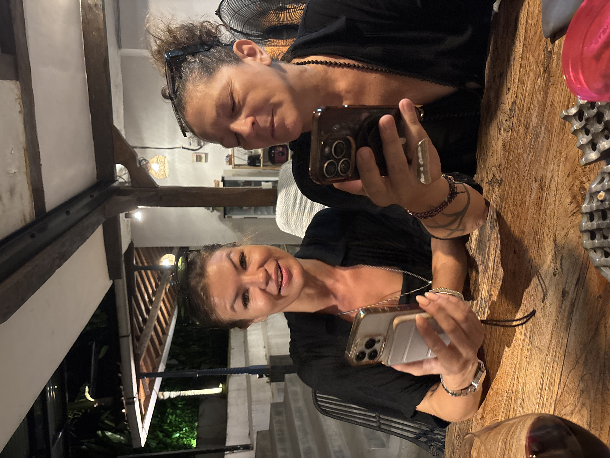 Two women are sitting at a wooden table, both holding smartphones. The woman on the left is looking at her phone, while the woman on the right is looking at the camera and smiling. Both women are wearing black shirts and jewelry. The table is made of rough wood and has a metal decoration on it. In the background, there are stairs, plants, and outdoor lighting, suggesting a location in North Kuta, Indonesia. The photo appears to be taken at a restaurant or outdoor venue.