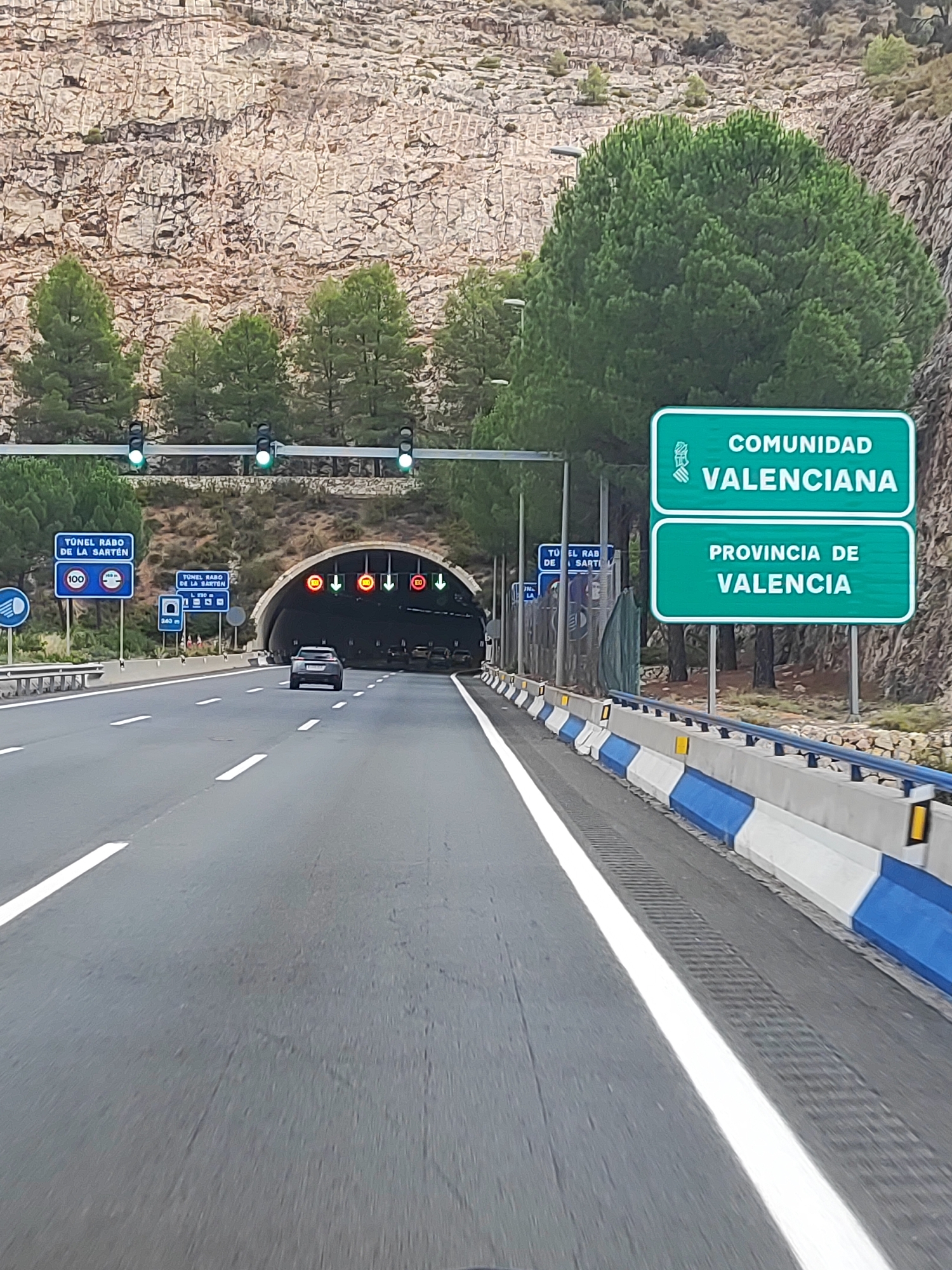 The image shows a highway approaching a tunnel. On the right side of the highway are two green signs, the top one reads "COMUNIDAD VALENCIANA" and the bottom one reads "PROVINCIA DE VALENCIA". These signs indicate that the driver is entering the Valencian Community and the Province of Valencia. The highway is paved and has white lane markings. A dark-colored car is driving on the highway, approaching the tunnel entrance. Several traffic lights are visible above the highway, some showing green. On the left side of the highway, there are blue signs indicating "TUNEL RABO DE LA SARTÉN" and a speed limit of 100. The tunnel entrance is wide and arched, and traffic lights are visible at the tunnel entrance. The surrounding environment includes a rocky hillside on the left and trees on the right, with a blue and white concrete barrier separating the highway from the roadside. The scene appears to be located in a mountainous or hilly region.