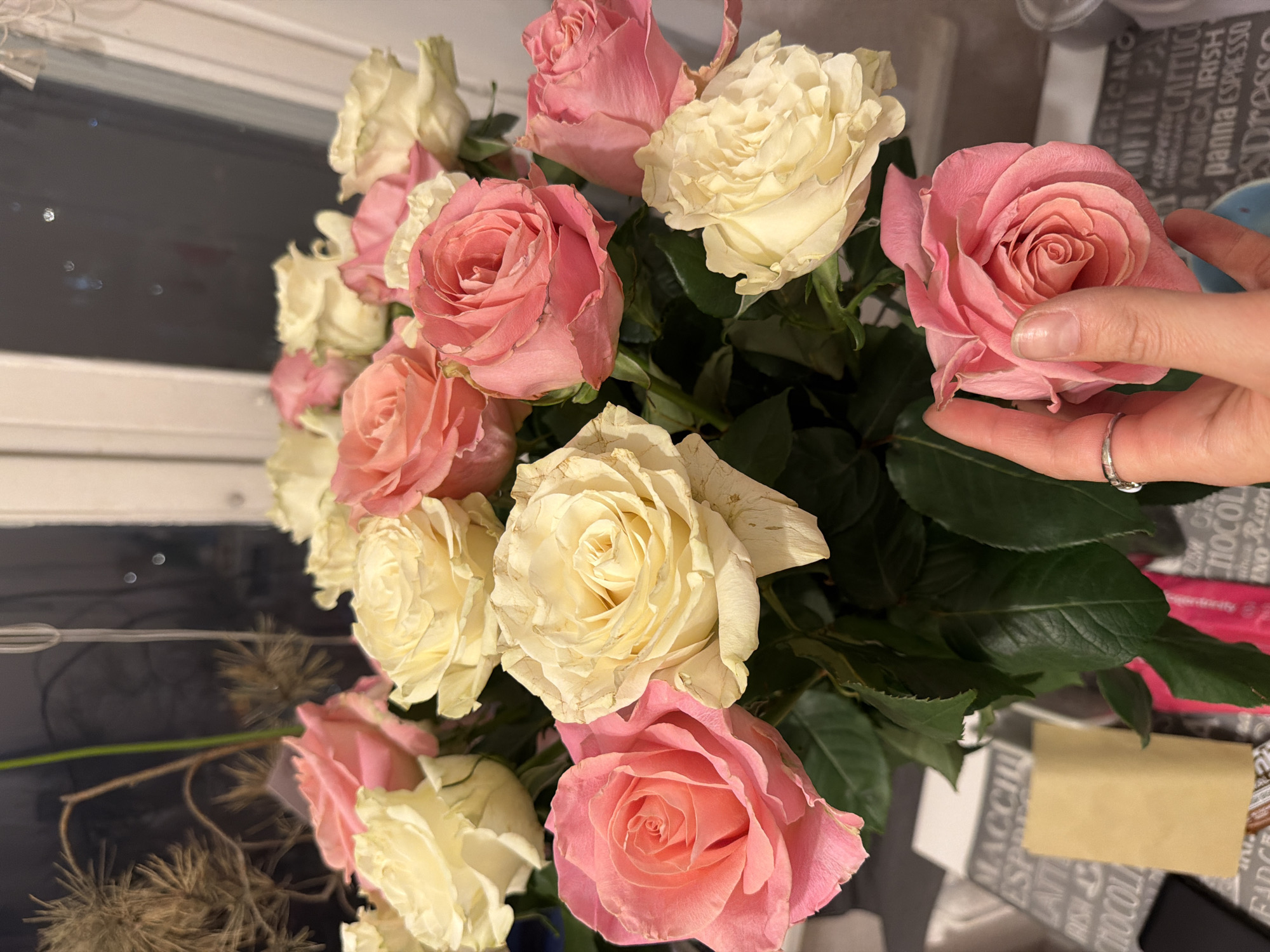 The image shows a bouquet of pink and white roses. A hand with a ring on one of the fingers is holding one of the pink roses. The bouquet is on a table with some items on it. The background is blurry but it looks like it could be a window or a wall. There is no indication of the location being in Moscow, Russia.