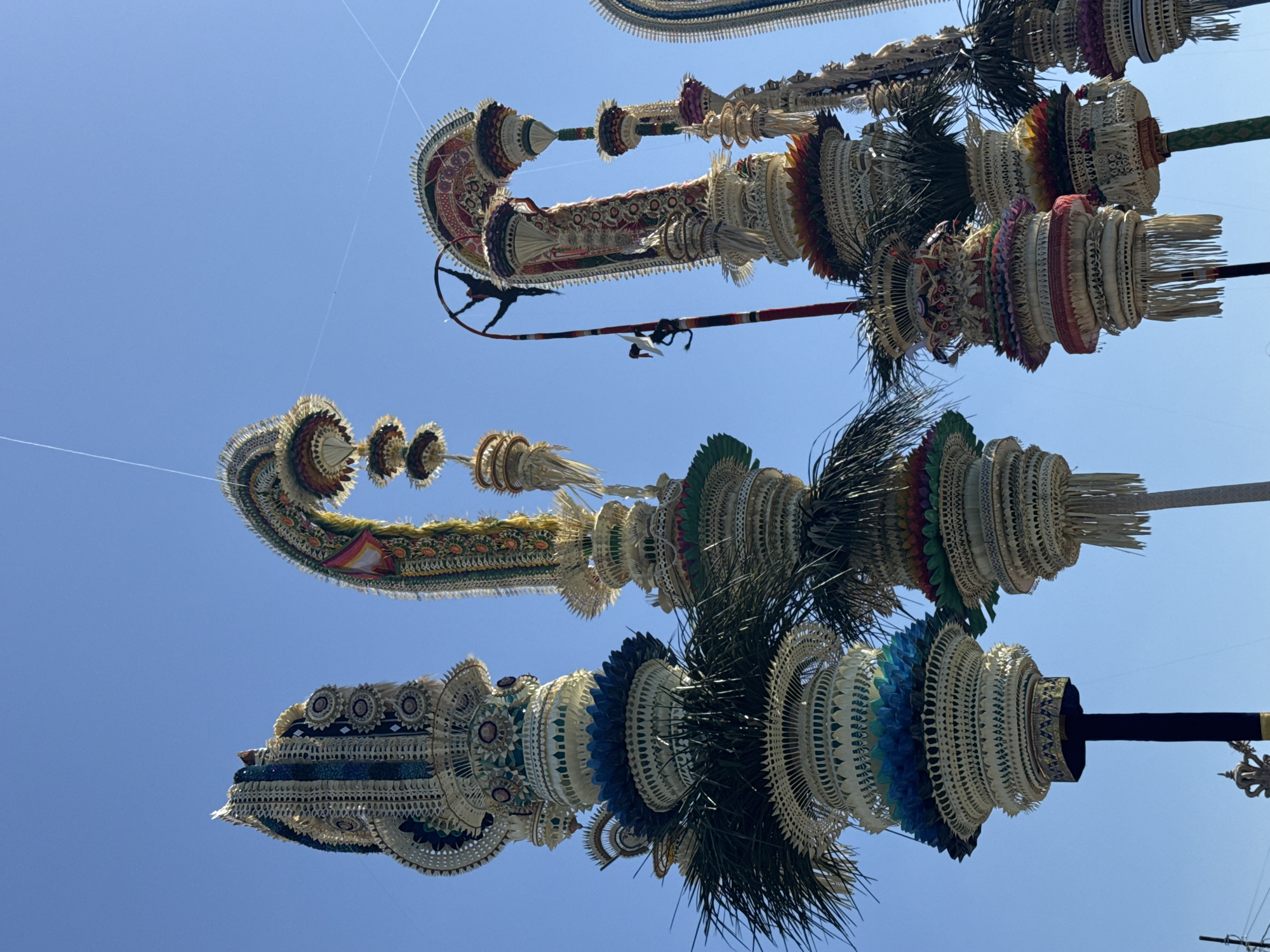 In Mengwi, Indonesia, a series of ornate, towering structures, likely part of a cultural or religious procession, are captured against a clear blue sky. These structures are elaborate, multi-tiered creations predominantly made from woven natural materials, such as palm leaves and straw, in shades of cream, white, and tan. They are adorned with vibrant, intricate detailing in colors like green, blue, red, and gold.
The structures vary in height and design, but many feature layered, circular elements resembling lotus flowers or headdresses. Some are topped with curved, arching elements that are also richly decorated. Green fronds, resembling palm leaves, extend outwards from some of the lower tiers. One of the structures has a dark pole, possibly a central support, wrapped in black fabric at its base. Another structure features a tall, slender pole with colorful banding and a dark, articulated figure or effigy attached to it, resembling a dancer or a mythological creature. This figure appears to be in a dynamic pose, with limbs extended. Fine white strings, possibly kite lines or decorative elements, are visible crisscrossing the sky above the structures. The overall impression is one of festive, elaborate craftsmanship and a vibrant outdoor event.