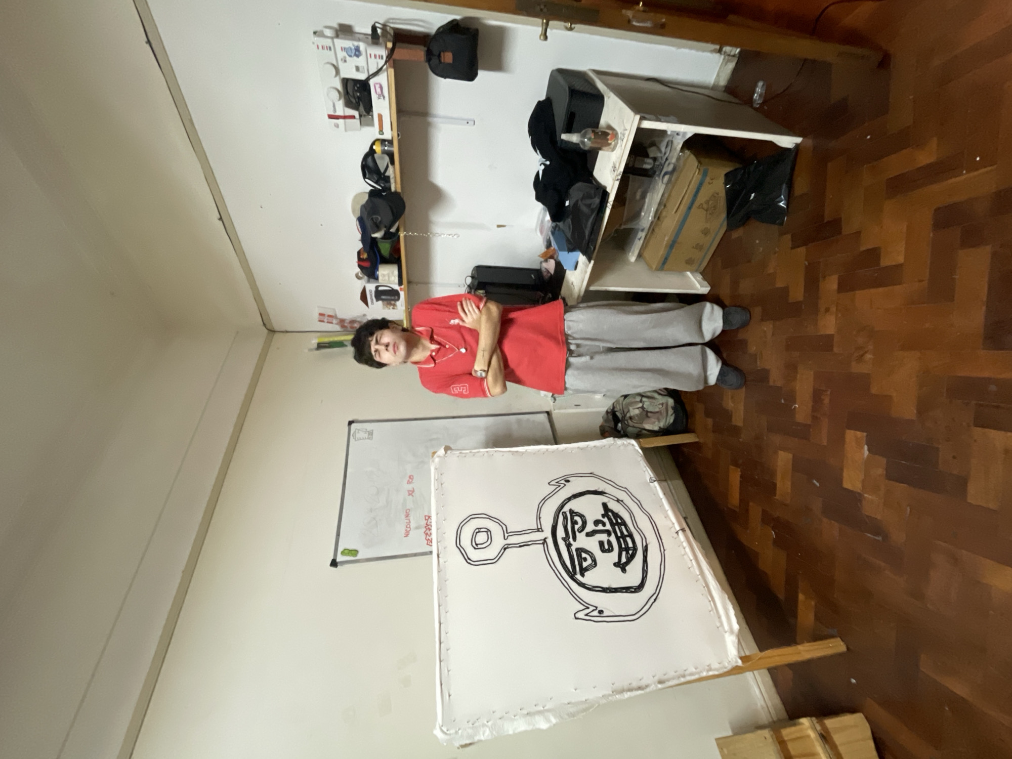 A person stands in a room with wooden parquet flooring and white walls. The person wears a red polo shirt and gray pants, and has their arms crossed. Behind them is a whiteboard and a piece of art propped up on a frame. There is a shelf cluttered with items on the wall, and a white desk with more items. The location is likely a home or studio. The city of Buenos Aires, Argentina, cannot be confirmed from the image.