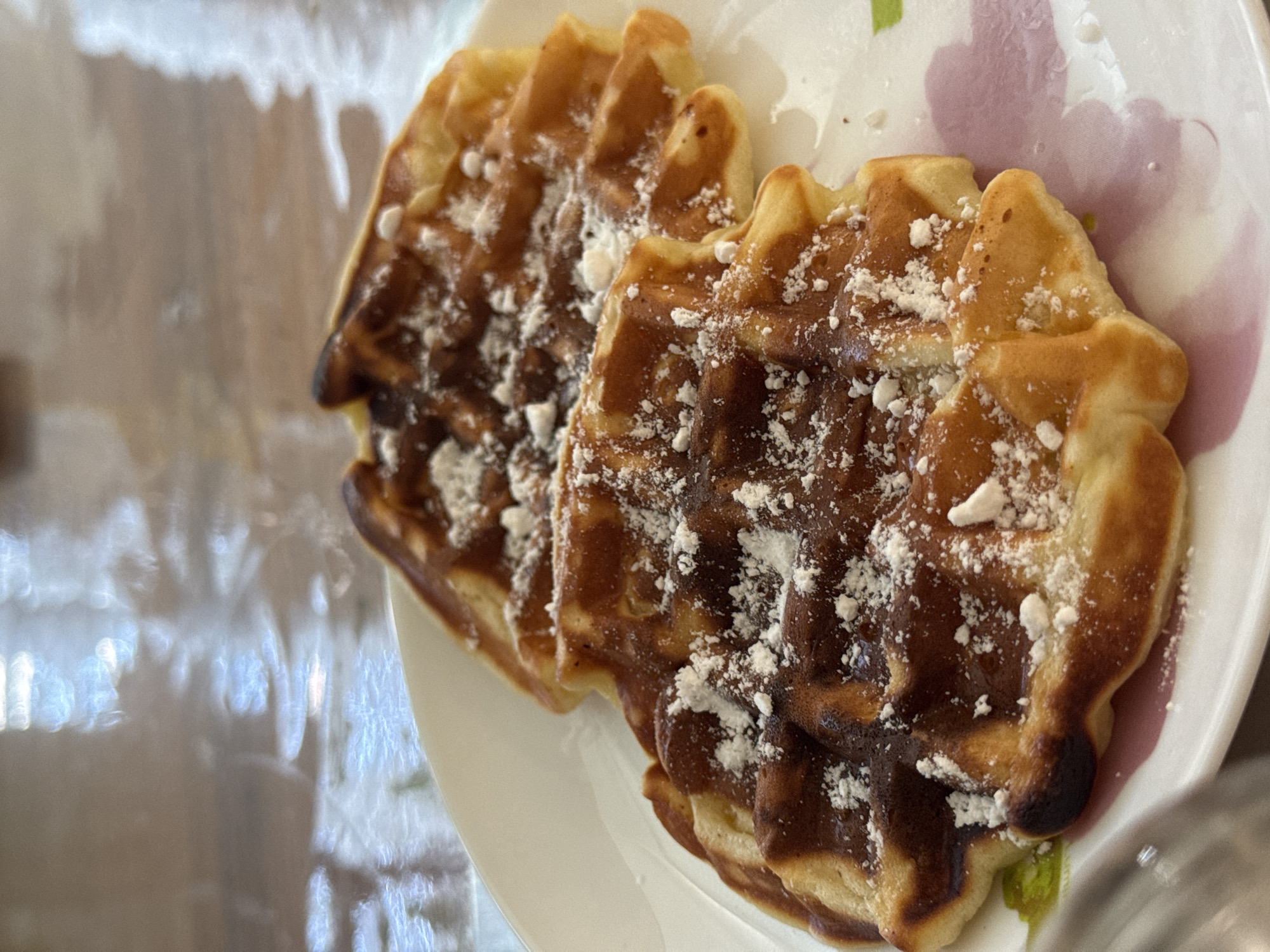 Two waffles dusted with powdered sugar sit on a white plate with a floral pattern. The waffles are golden brown and have a slightly irregular, scalloped shape. The background appears to be a reflective, possibly glass-covered, surface. There is no indication of the location being Chaikovsky, Russia.