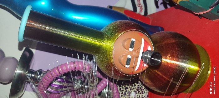A whimsical, horizontally positioned object with a brown, cartoonish face is displayed. The face features prominent white-outlined eyes and red "lips." This is affixed to a reflective, gradient cylinder transitioning from yellow to brown. Delicate fibers radiate from the base of the "face." Other assorted trinkets and a portion of a blue object are in the background. A turquoise ring encircles the cylinder.