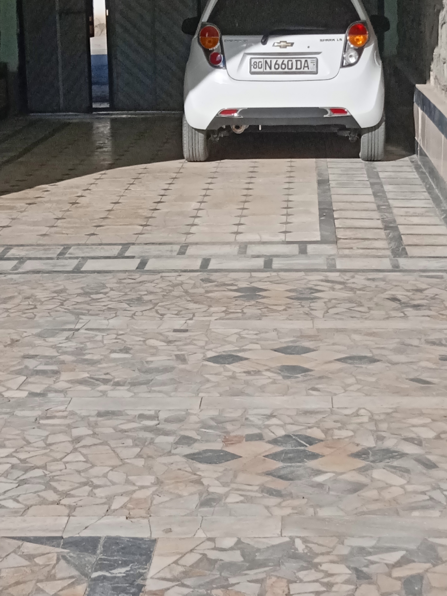 In an outdoor setting, the rear of a white Chevrolet Spark LS is parked on a tiled surface. The car's license plate reads "8G N660 DA". To the left, large metal garage doors are partially open, revealing a dark interior. The tiled ground features a geometric pattern with rectangular and square tiles, bordered by a strip of larger rectangular tiles. Further away, the tiling shifts to a mosaic of irregularly shaped stones in various shades of gray, beige, and black, forming diamond-shaped patterns. The lighting suggests it is either dusk or dawn, with long shadows cast across the ground from the open garage.