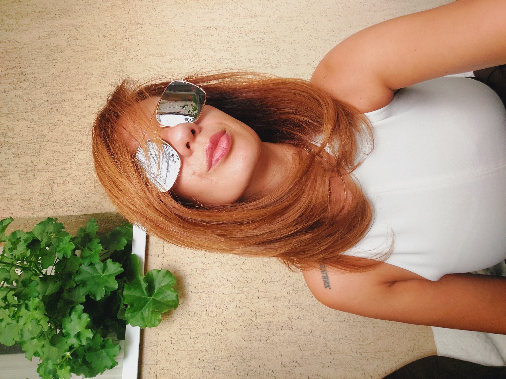 A woman with auburn hair and sunglasses is posing for a selfie in front of a textured beige wall. She is wearing a white sleeveless top and a delicate gold necklace. A green plant is visible in the upper left corner of the image, and a tattoo of Roman numerals is on her left shoulder. The reflection in her sunglasses shows a potted plant with pink flowers. The scene appears to be outdoors, possibly on a balcony or patio. The image was captured in Sofia, Bulgaria.