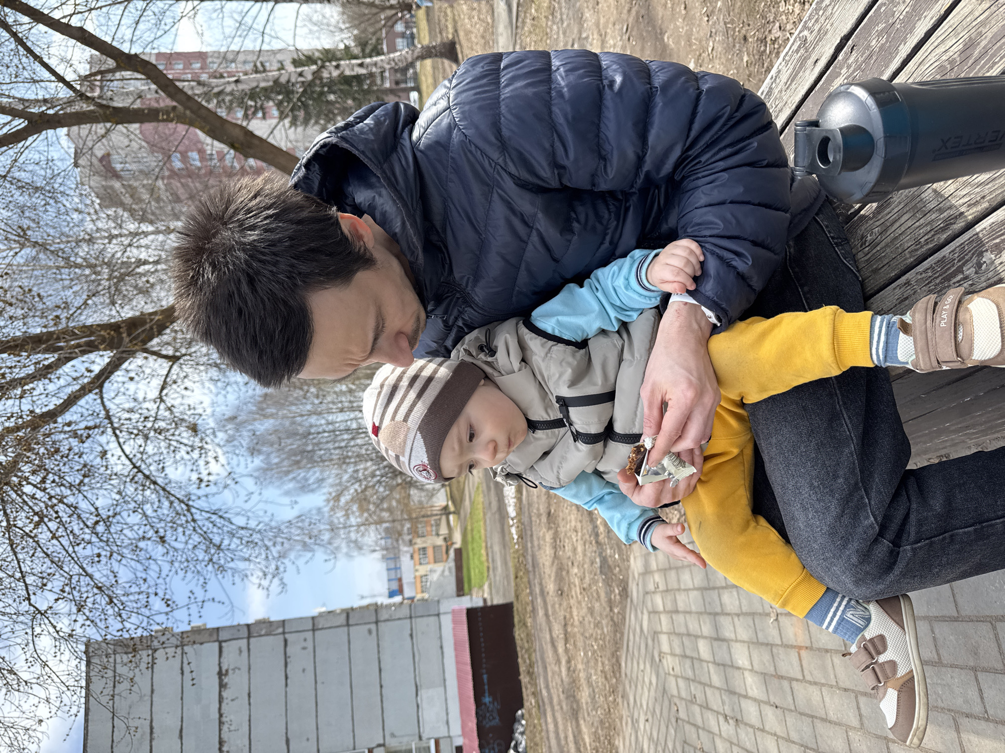 A medium shot depicts an adult male seated on a wooden park bench, cradling a toddler on his lap. The man, clad in a navy puffer jacket and dark jeans, leans over the child, whose attention is drawn to a piece of food or candy being held in the man's hand. The toddler is dressed in a striped beanie, a light gray vest, a light blue undershirt, and yellow pants. The child wears sneakers with light-colored socks that have dark blue stripes.
The background includes bare trees, a brick-paved ground, and a multi-story building typical of urban landscapes in Izhevsk, Russia. A large black water bottle sits behind the man on the bench. Graffiti is visible on a concrete wall.