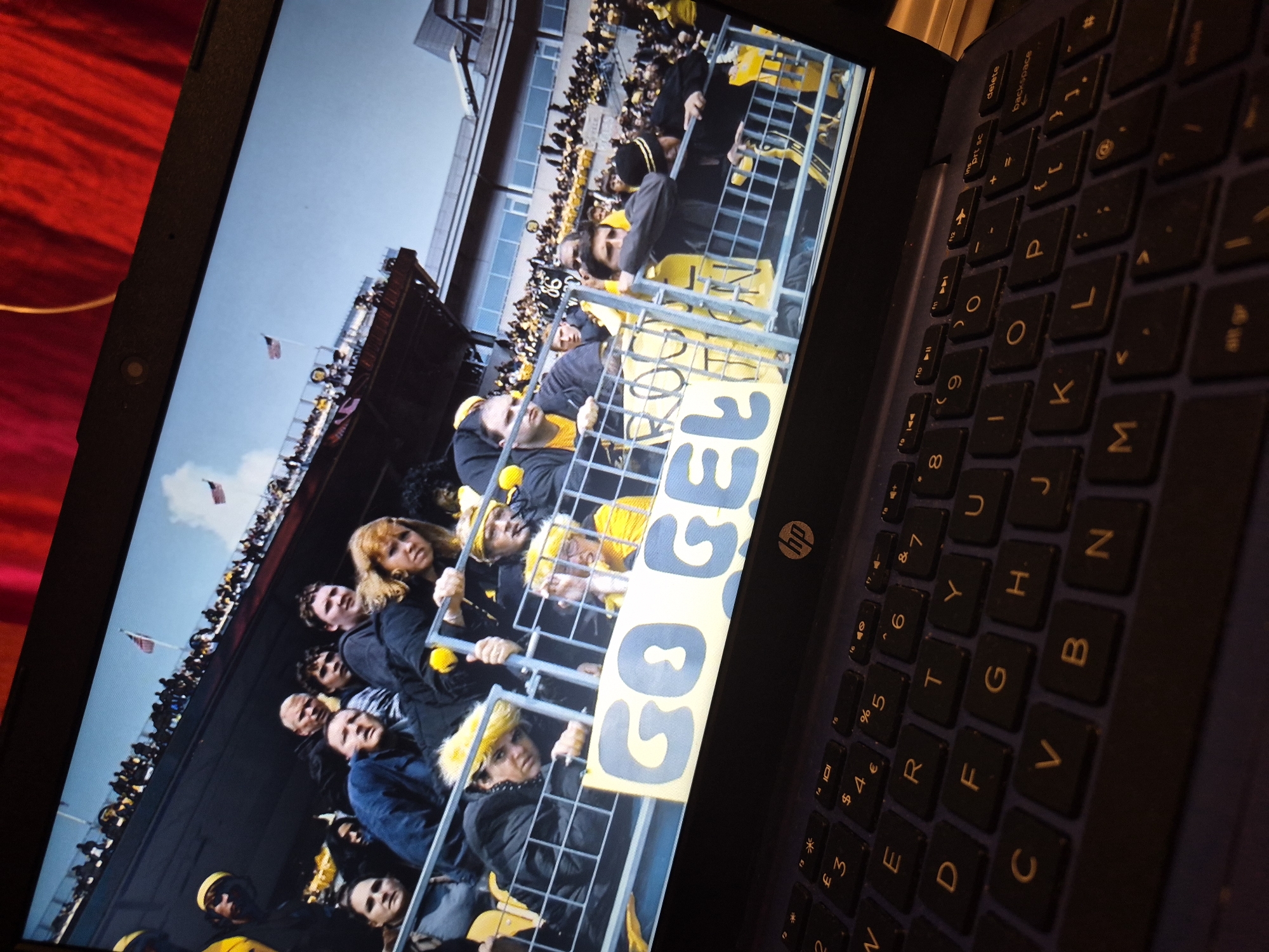 The image shows a laptop screen displaying a photograph of a crowd of people in what appears to be a stadium or arena. The people are mostly wearing black and yellow, suggesting they are fans of a sports team. Some are holding a banner that says "GO GE...". In the foreground, the keyboard of the laptop is visible, with keys such as 'V', 'B', 'N', 'G', 'H', 'J', 'K', 'L', 'M', 'O', 'P', 'I', 'U', 'Y', 'T', 'R', 'E', 'W', 'Q', 'A', 'S', 'D', 'F' visible. The background of the laptop screen shows a red curtain or fabric. The overall scene depicts a person viewing a sporting event on their laptop. There is no information to suggest the location is in Sandwell, United Kingdom.