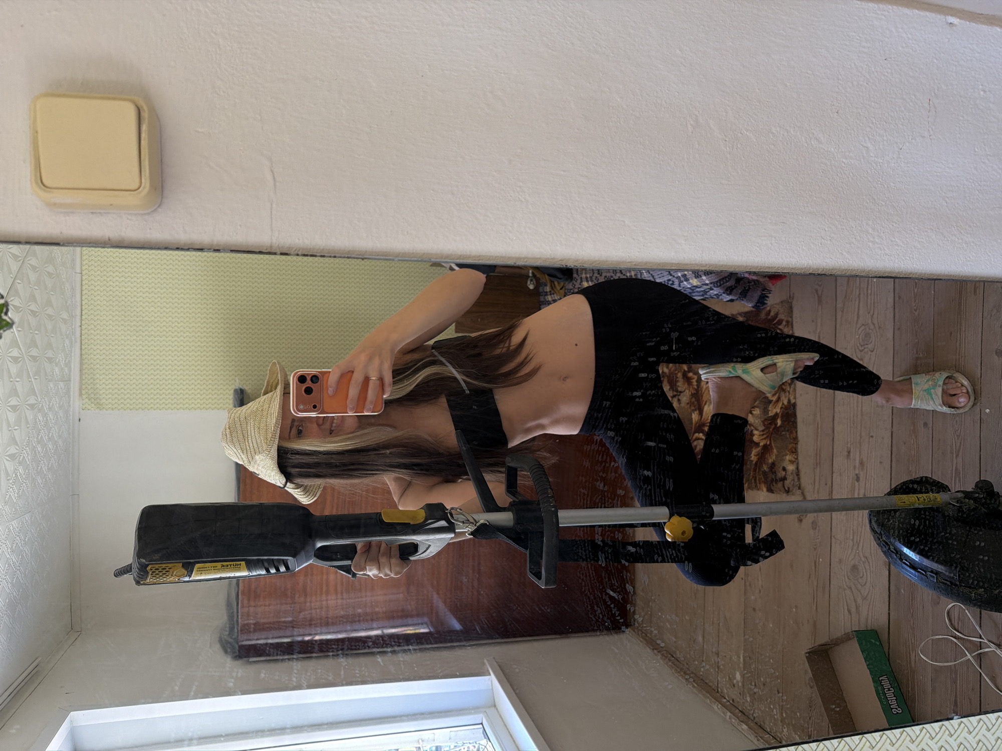 A reflection of a woman holding a weed whacker is seen in a mirror. The woman is wearing a black top, black leggings, and a straw hat. She is holding a phone in her left hand, taking a selfie in the mirror. The weed whacker is black and yellow. The environment appears to be a room with white walls and wooden floors. Based on the visual elements, it's not possible to confirm the location in Kamyshin, Russia.