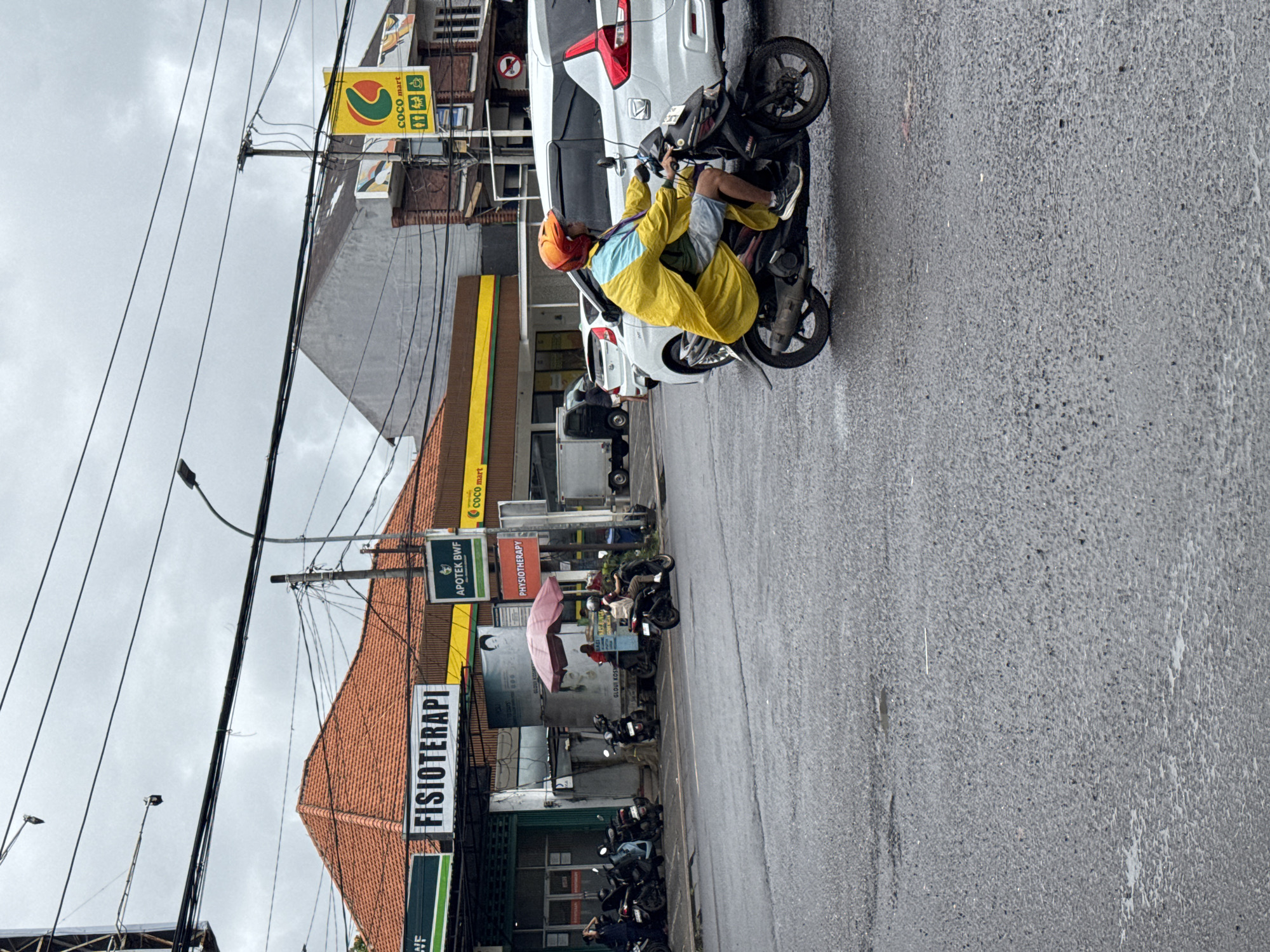 This image captures a daytime street scene in Kuta Selatan, Indonesia, under an overcast sky. The asphalt road appears wet, suggesting recent rain.
In the foreground on the right side of the street, a person wearing a bright yellow rain poncho and an orange helmet is seated on a black scooter, parked next to a white car. The vehicles are facing away from the viewer.
Along the left side of the street, several buildings line the thoroughfare. One building features a distinctive reddish-brown tiled roof and a prominent sign reading "FISIOTERAPI" (Physiotherapy). Above it, another sign displays "APOTEK BWF" (BWF Pharmacy). Further down, a bright yellow sign with a green logo is visible, indicating a "COCO mart." A large poster with a person's face is also visible on one of the buildings. Several other scooters are parked along the sidewalk, and a pink umbrella provides shelter near what appears to be a small street-side setup.
Above the street and buildings, numerous utility poles are interconnected by a dense web of black power lines, indicative of an urban or semi-urban environment. The scene depicts typical daily life on a bustling street, with commercial establishments and various modes of transport, during potentially wet weather conditions.