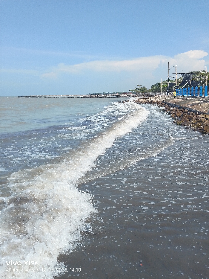 The image displays a coastal scene in Limbangan, Indonesia, captured on October 12, 2025, at 14:10. The foreground features murky, greyish-brown ocean water with multiple white-capped waves actively breaking towards a dark, sandy or muddy shoreline. To the right, a rocky embankment or seawall forms the immediate edge of the land. Beyond this rock barrier, an elevated area supports various structures, including vertical poles, dark tarpaulin or canvas coverings indicative of shelters or stalls, and several distinct blue, vertical fence-like barriers. Green foliage and trees are visible behind these structures. In the midground, extending into the water on the left, a long, low, stone breakwater or jetty is present. The horizon shows distant land features or structures. The sky is clear blue with sparse white stratocumulus clouds. No individuals are visible in the scene.