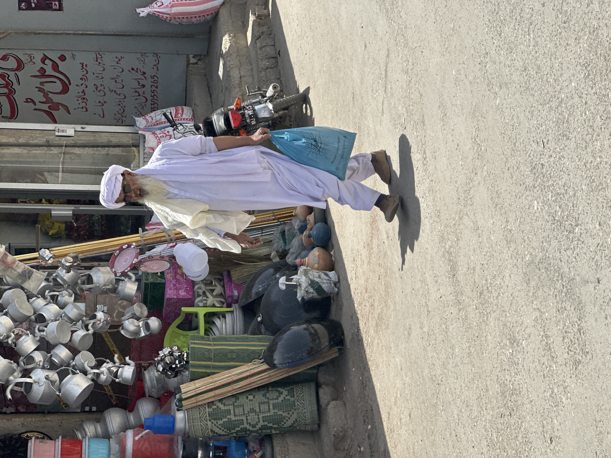 In the city of Kanozai, Pakistan, an elderly man walks along a dusty street in front of a shop. He is dressed in traditional white attire, including a turban and a long robe, and sports a prominent white beard and glasses. He carries a bright blue plastic bag in his right hand, which displays the words "Thank You" and "Please Come Again" in English. The shop behind him is adorned with a variety of metal pots, pans, and kitchenware hanging from its facade, along with stacks of containers and other goods. A motorcycle is parked near the shop entrance. The ground is paved with asphalt, and shadows from the man and other unseen objects are cast across it.