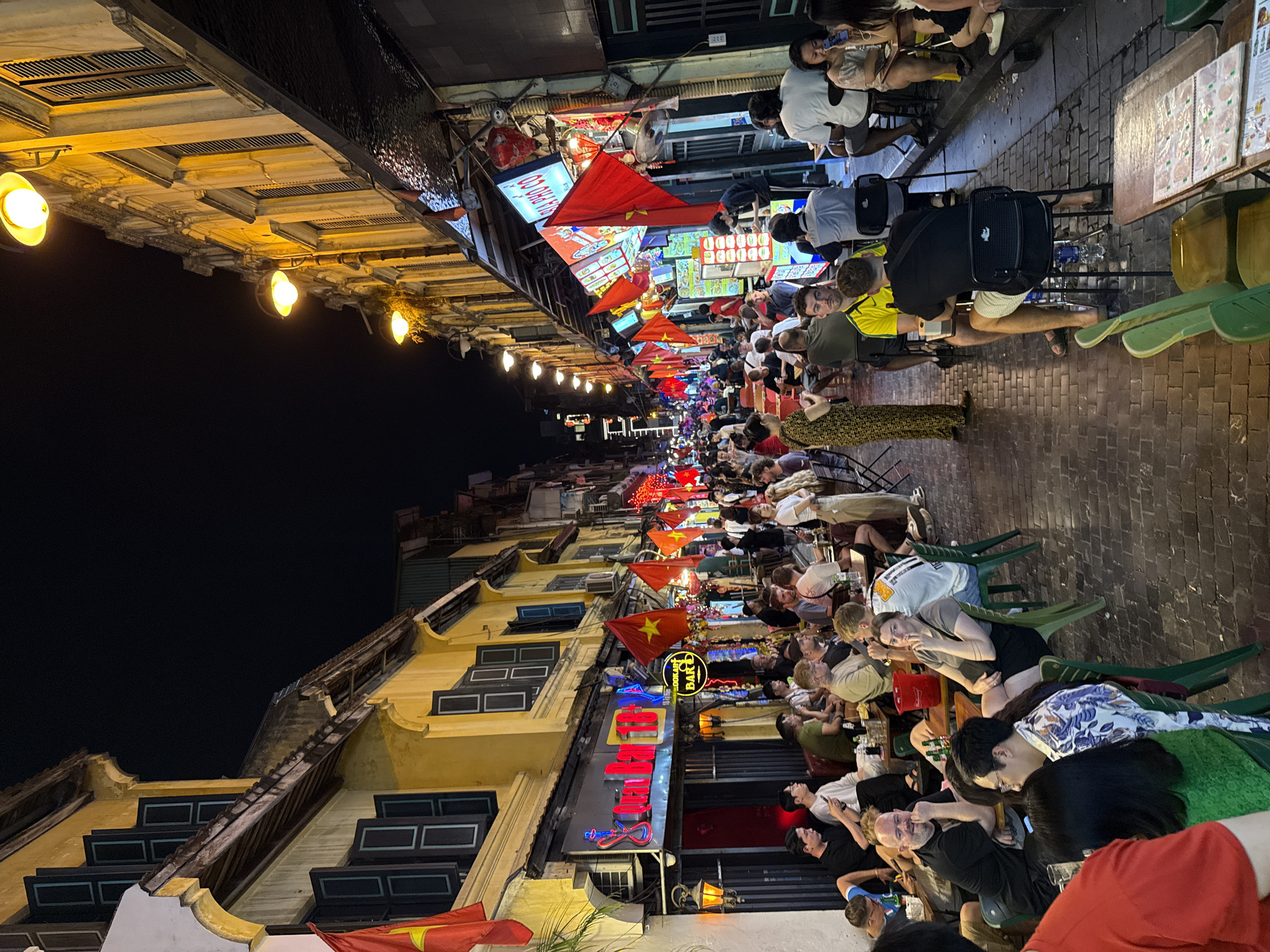 This image depicts a bustling night scene on a lively street in Hà Nội, Vietnam. Numerous people are gathered outdoors, seated at small plastic tables and chairs that line the brick-paved sidewalks. They appear to be socializing, dining, and drinking, contributing to a convivial atmosphere.
The time of day is night, with the dark sky contrasting with the warm, artificial lighting from streetlights, shop signs, and overhead bulbs. Old buildings with characteristic yellow facades and dark-shuttered windows run along the street, alongside various shopfronts.
A prominent feature throughout the scene is the abundance of red flags with a yellow star, the national flag of Vietnam, which are strung across the street or displayed on establishments. Other visible objects include menu boards, water bottles on tables, and a black backpack.
Textual cues on signs include "QUÁN BIA" (Beer Pub) and "HOKKAH BAR" (likely Hookah Bar), indicating the types of establishments present. The casual attire of the people and the crowded, informal outdoor dining setup are typical of street culture in Hà Nội, particularly in its Old Quarter. The overall impression is one of a vibrant and popular evening gathering spot.