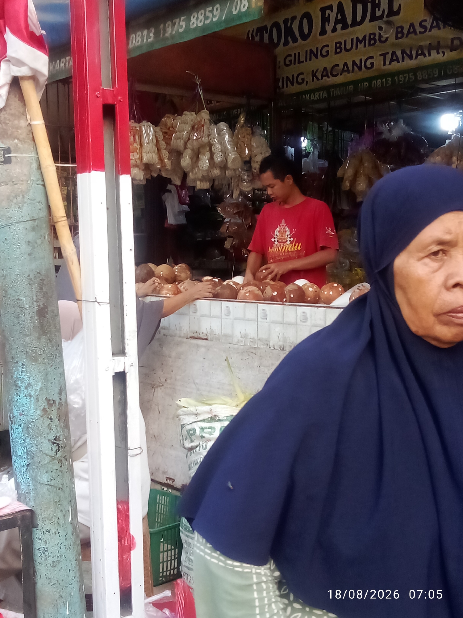 The image depicts a street-side market stall in East Jakarta, Indonesia. In the foreground, a woman wearing a dark blue hijab is partially visible, looking towards the right side of the frame. Behind her, a young man in a red t-shirt is standing behind a counter, arranging coconuts. The stall is filled with various goods, including bags of dried snacks hanging from the ceiling and piles of coconuts on display. A sign above the stall reads "TOKO FADEL: GILING BUMBU BASA". The environment appears to be an outdoor market, with a red and white striped pole to the left of the frame and a distressed blue metal pole behind it. The timestamp "18/08/2026 07:05" is visible in the bottom right corner.