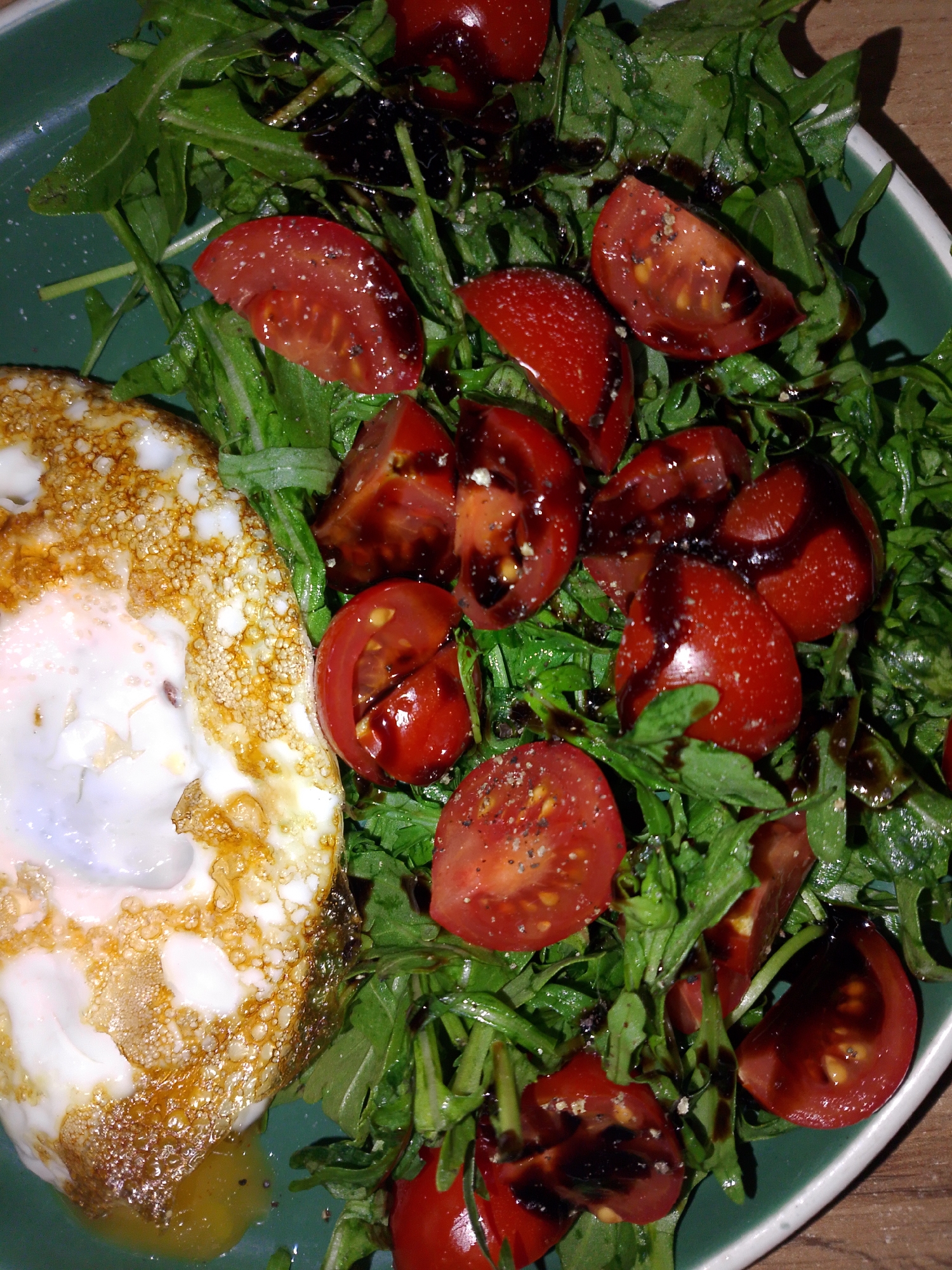A close-up shot displays a plate of food. The plate contains a fried egg, a bed of arugula salad, and halved cherry tomatoes drizzled with balsamic glaze. The egg is fried with crispy edges. The arugula is fresh and green. The cherry tomatoes are red and succulent. The balsamic glaze adds a dark, glossy finish to the tomatoes and is also present on the greens. The plate appears to be green with a white rim. The background is out of focus, revealing a wooden surface. There is no information to suggest the location is Izhevsk, Russia.
