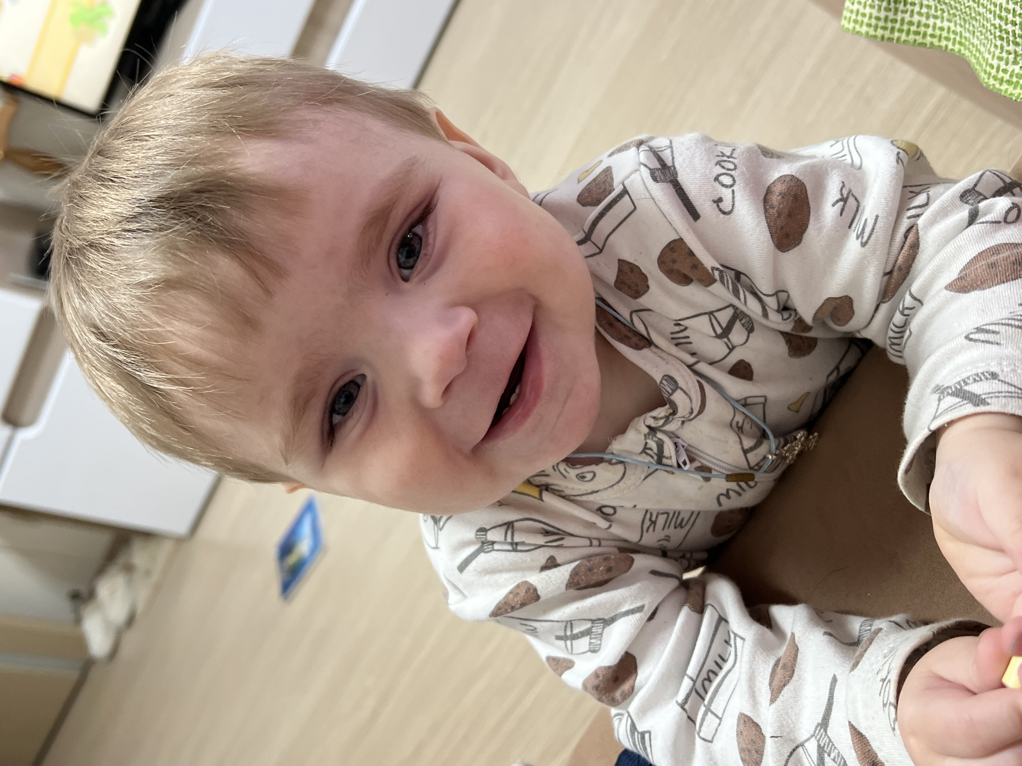 A young child is shown close-up, smiling and looking up at the camera. The child has light blond hair and blue eyes. The child is wearing a white pajama onesie with a repeating pattern of milk cartons, cookies, and baking utensils. The child is laying on a brown surface, possibly a person's lap. In the background, there is a wooden floor, a white cabinet, and a green patterned cloth. The image was taken indoors and is well-lit. There is no information provided in the image to confirm the location being the city of Perm, Russia.