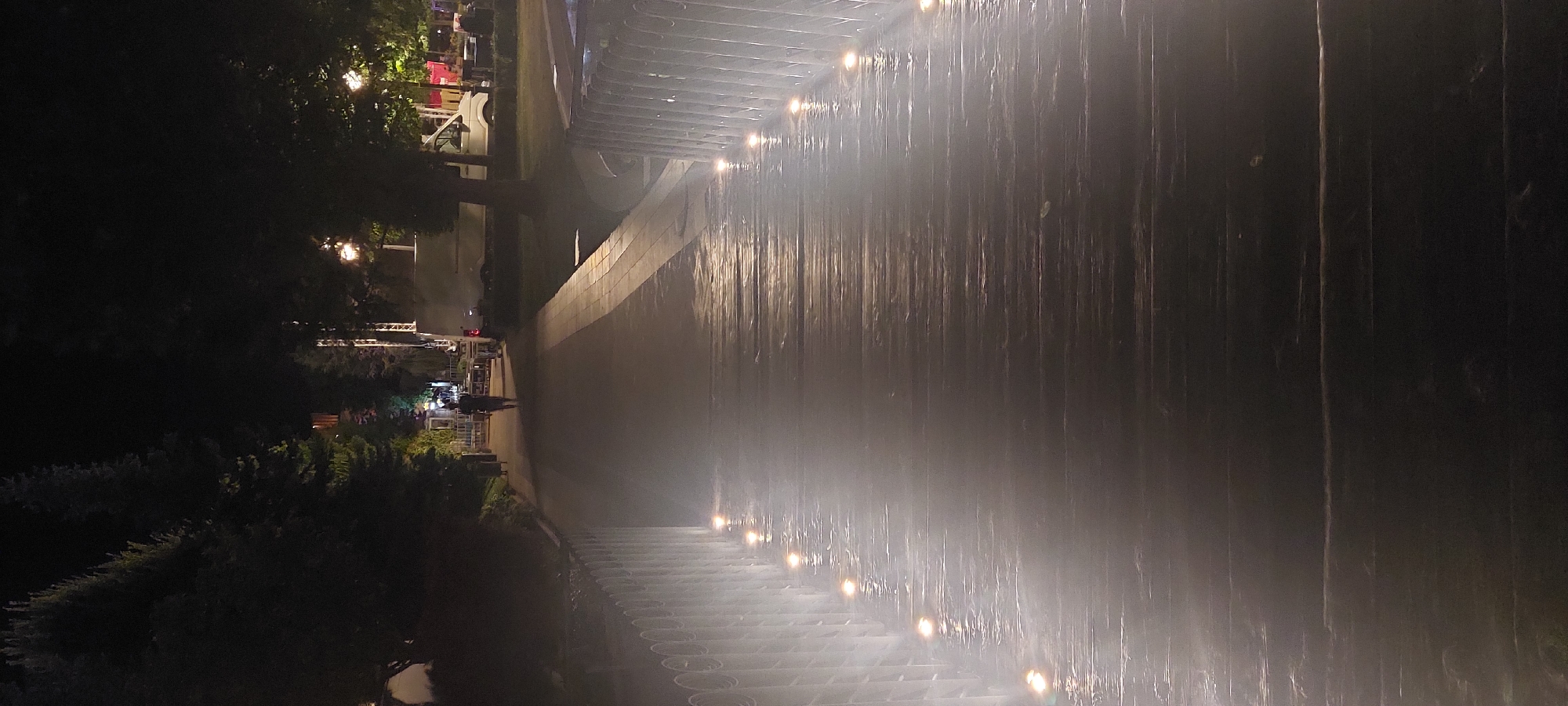 The image depicts a nighttime scene of a pedestrian bridge, likely in a park or public area in Iași, Romania. The bridge has a wooden deck illuminated by numerous small, embedded lights along its sides, creating a foggy or misty effect due to the low-angle lighting and possibly moisture. A solitary figure, dressed in dark clothing, is walking away from the camera towards the far end of the bridge. The background shows trees, some streetlights, and what appears to be a temporary setup with barriers and a white van, suggesting an event or outdoor market. The overall atmosphere is dark and somewhat atmospheric, with the lighting on the bridge being a prominent feature.