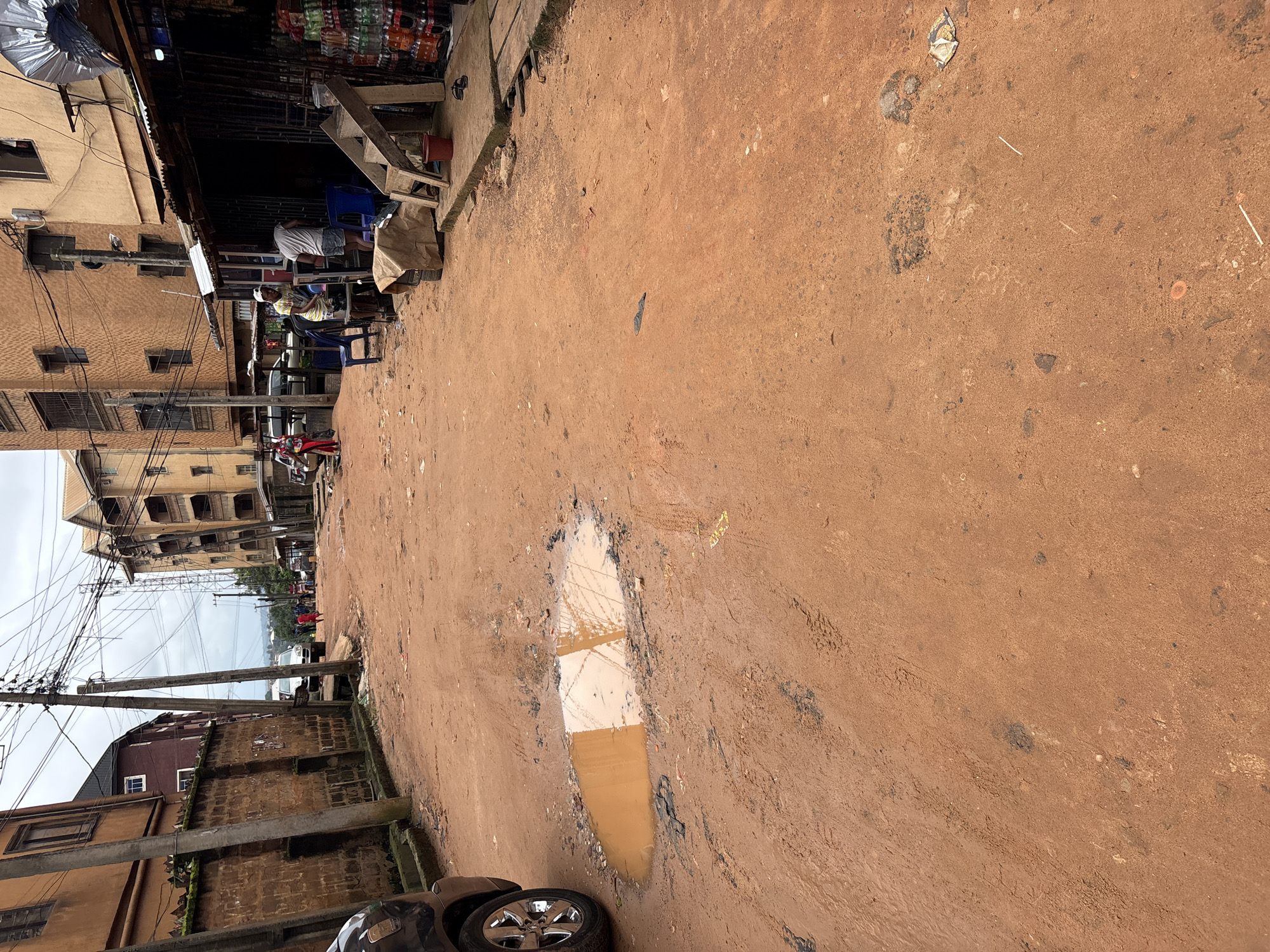 The image depicts a street in Nkpor, Nigeria, characterized by its unpaved, reddish-brown earth surface and the presence of several buildings. The street appears to be a thoroughfare with some traffic, as indicated by the vehicles visible. On the right side of the street, a shop is set up with various goods, including bottled drinks displayed on shelves. Several people are present, some of whom appear to be customers or vendors associated with the shop. There is a puddle of murky water on the left side of the road, suggesting recent rainfall or inadequate drainage. The environment appears to be a typical urban street scene in a developing area, with overhead power lines and a mix of residential and commercial structures. The overall impression is one of a busy, functional street that reflects the local economic and social activities.