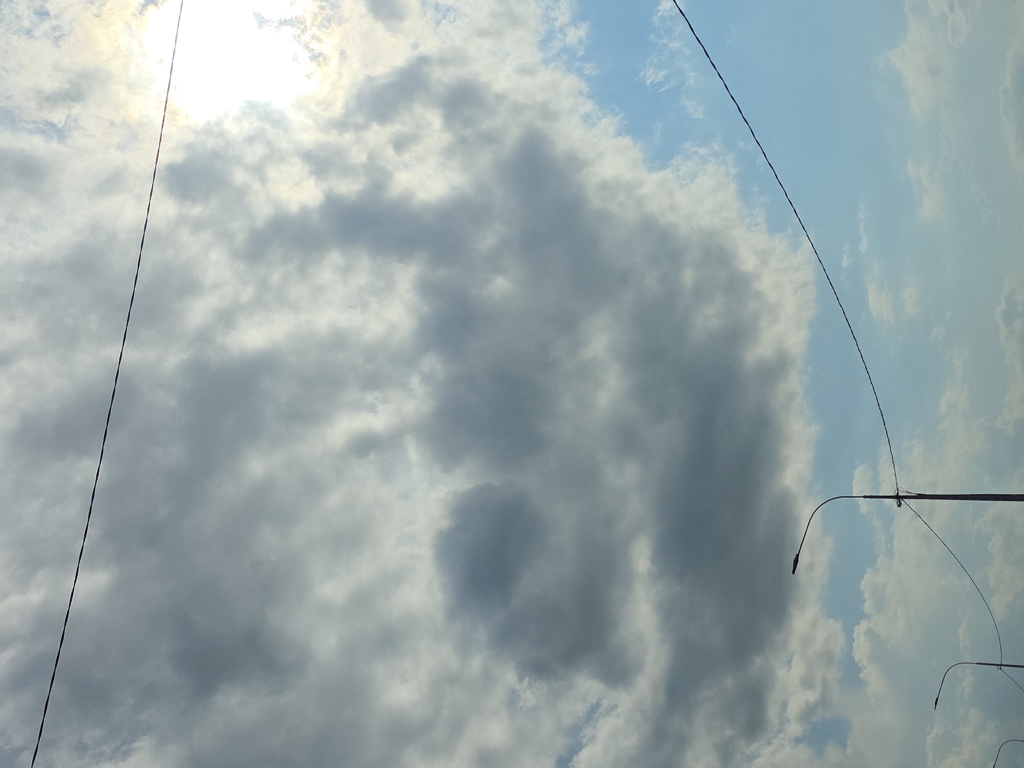 The image depicts a cloudy sky with a bright sun peeking through on the right side. Two streetlights with curved arms are visible in the lower portion of the frame, along with wires stretching across the sky. The scene appears to be an outdoor urban environment.