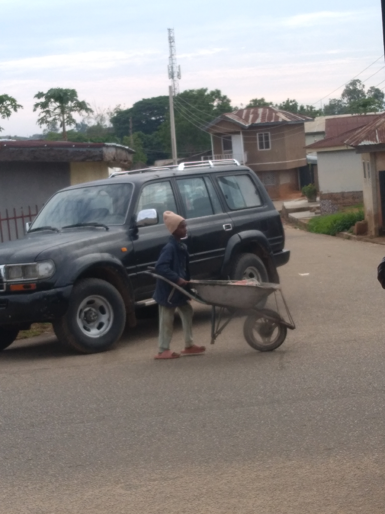 The image depicts an outdoor street scene within a semi-urban or rural area of the city of Unknown, Unknown, under an overcast sky. A young individual, seen from head to toe, wears a light brown beanie, a dark blue long-sleeved garment, light beige trousers, and reddish-brown open-toed sandals. This individual is actively pushing a grey metallic wheelbarrow, which contains light-colored material, along a paved asphalt road. To the immediate left of the individual and behind the wheelbarrow, a black Toyota Land Cruiser 80 series SUV, equipped with a roof rack, is parked facing left on the roadside. The vehicle's front quarter panel, headlight, and front wheel are clearly visible. In the background, on the left, a building with a dark roof and a red metal fence is partially visible. Further into the background, several residential structures with light-colored walls and distinct rusty corrugated metal roofs are present, along with dense green foliage and various trees. A tall metal communication tower stands prominently on the right side of the background, and utility wires are visible against the sky.