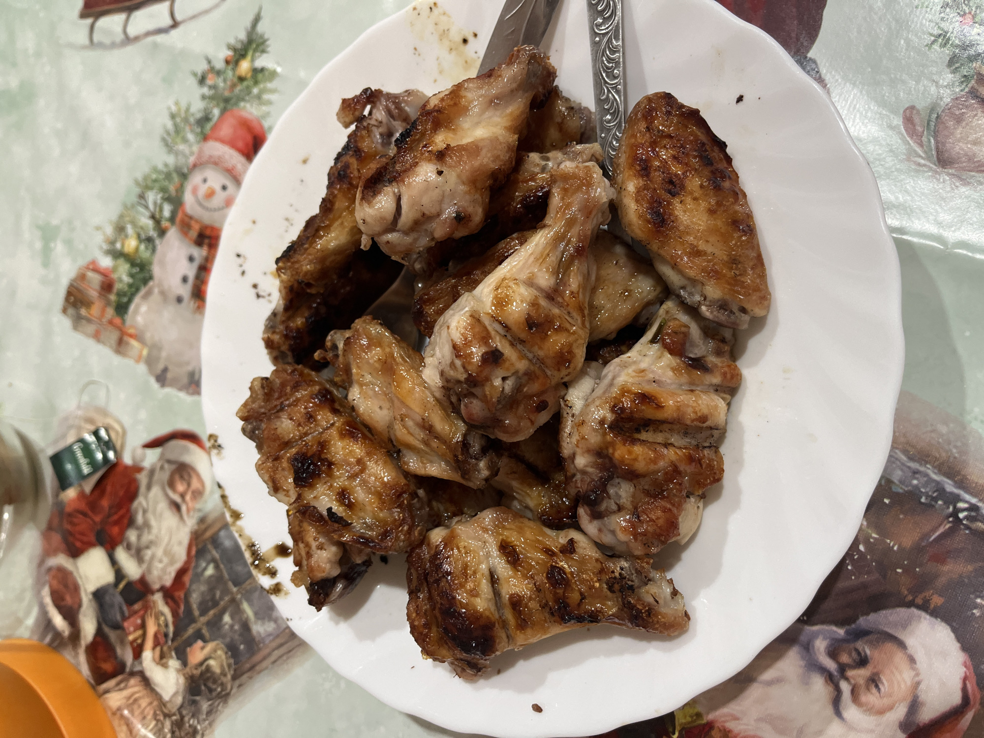 A white plate filled with cooked chicken wings sits on a tablecloth decorated with Christmas-themed designs, including a snowman and Santa Claus. A fork and a knife are partially visible amidst the chicken wings. The location is Chaikovsky, Russia.